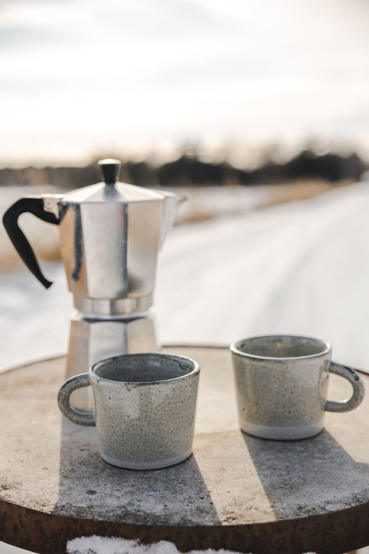 A moka pot coffee maker sits next to a speckled gray ceramic coffee mug on a textured stone surface.