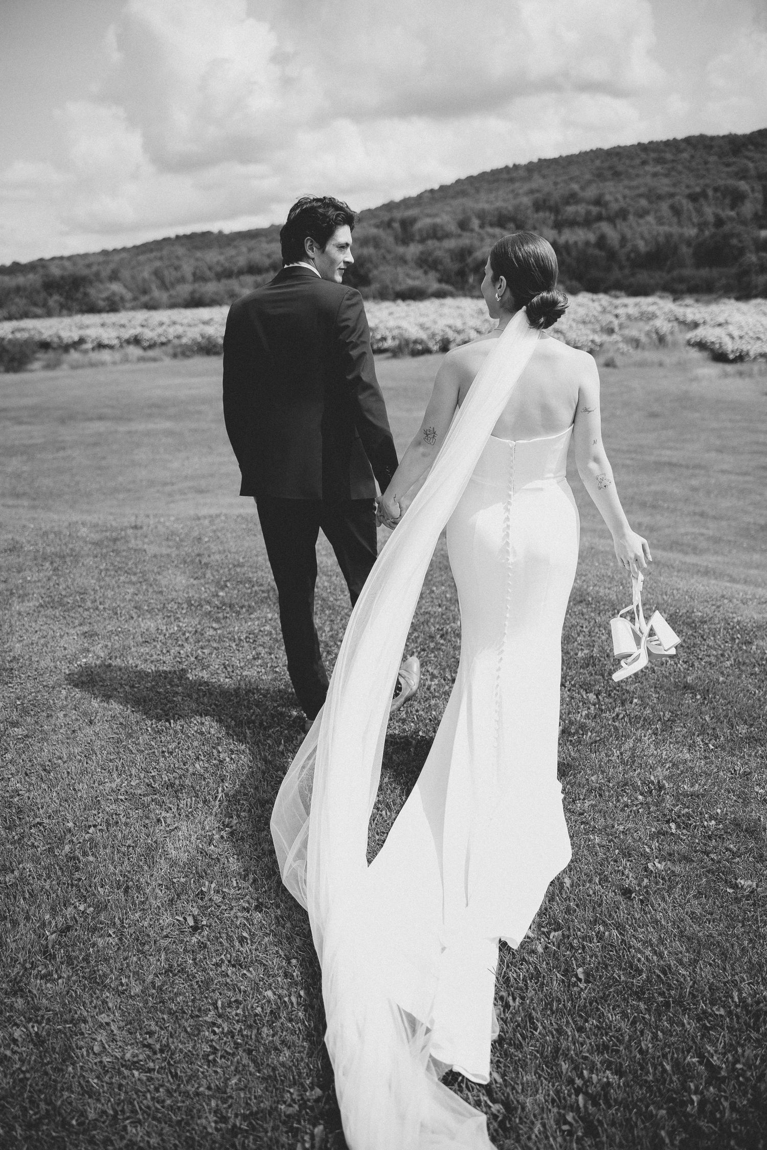 Black and white photo of a couple walking through a grassy field in wedding attire.