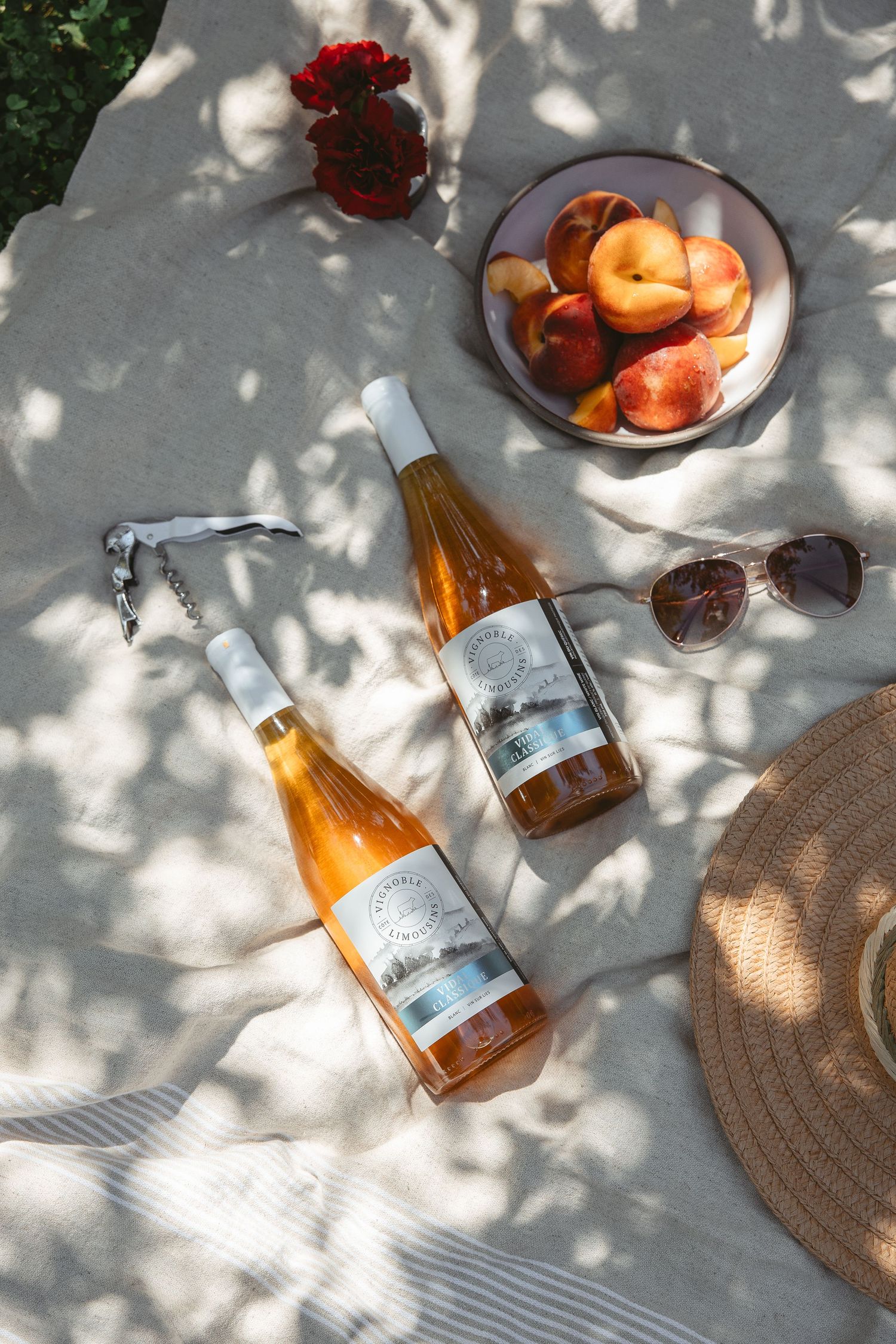 Overhead view of orange wine bottles and peaches on a sunlit surface with dappled shadows and a woven placemat.
