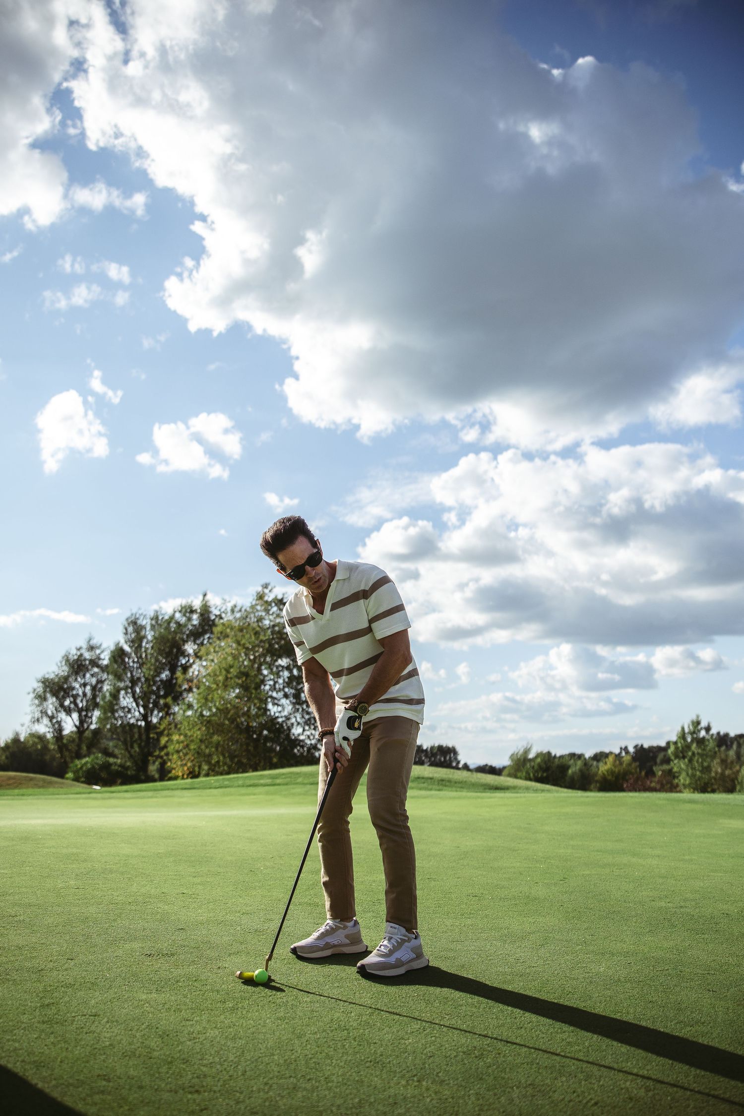 Golfer lines up a putt on a sunny golf course with dramatic clouds in the background.