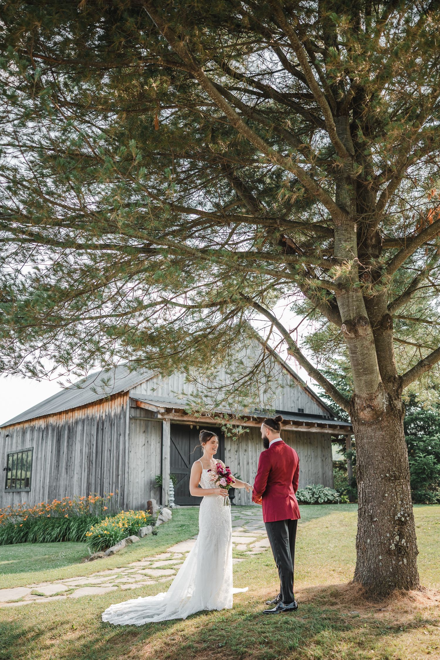 Couple de mariés debout sous un grand pin, près d’une grange rustique en bois, lors d’un mariage champêtre en pleine nature.