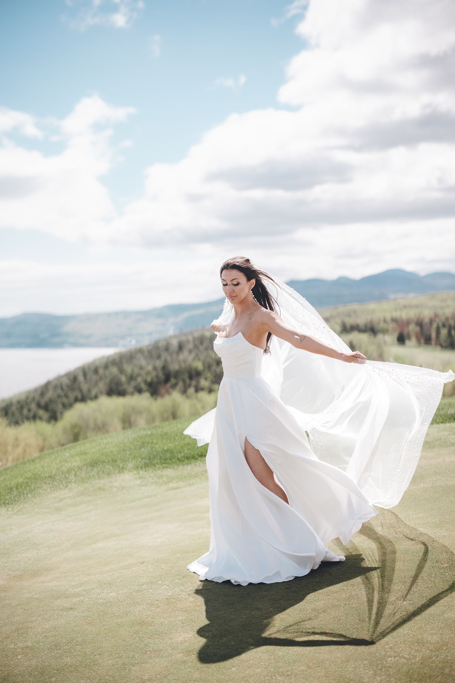 Une robe de mariée blanche et fluide flotte au vent, devant un paysage montagneux spectaculaire.