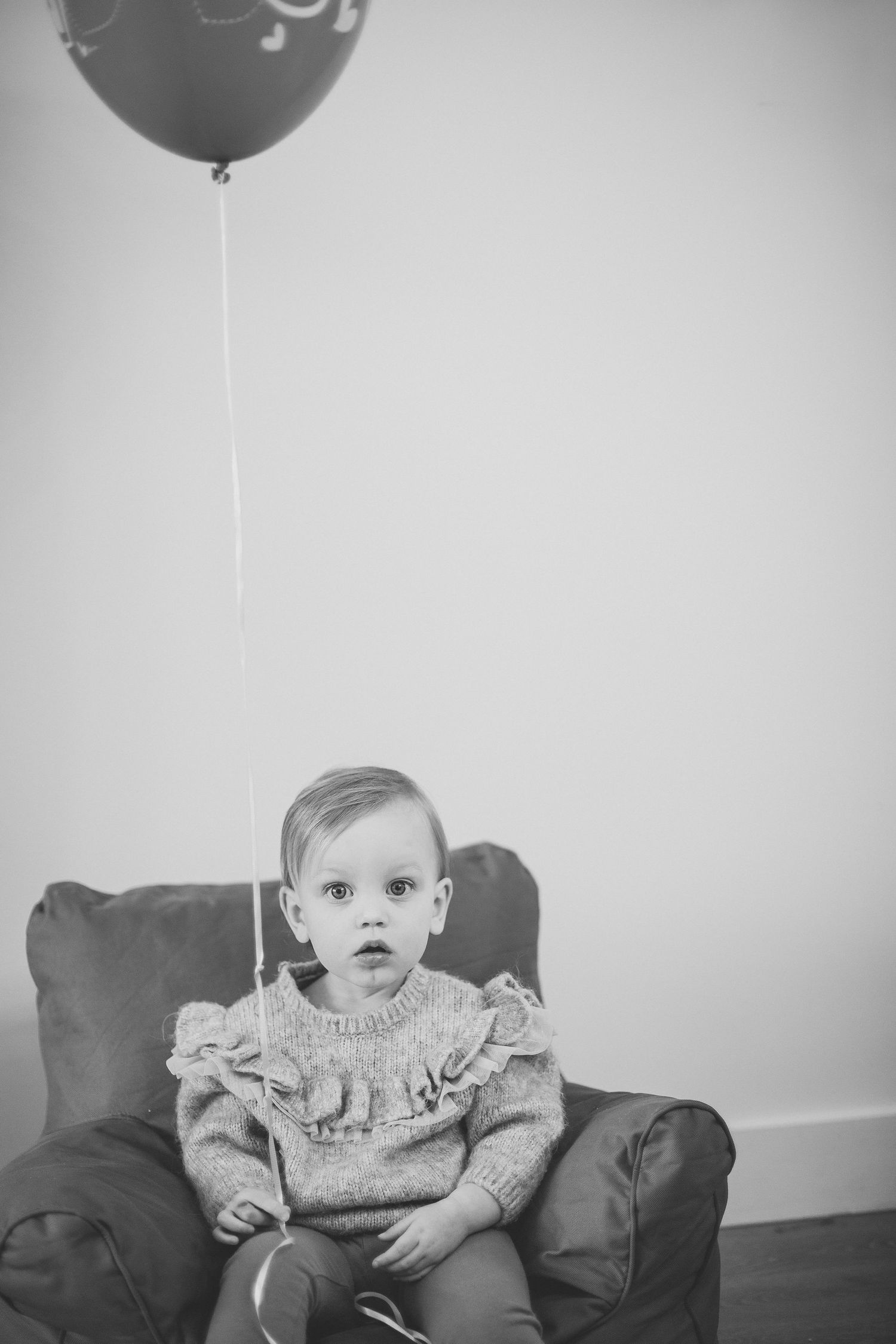 A baby sits on a gray couch with a floating balloon in a minimalist setting.