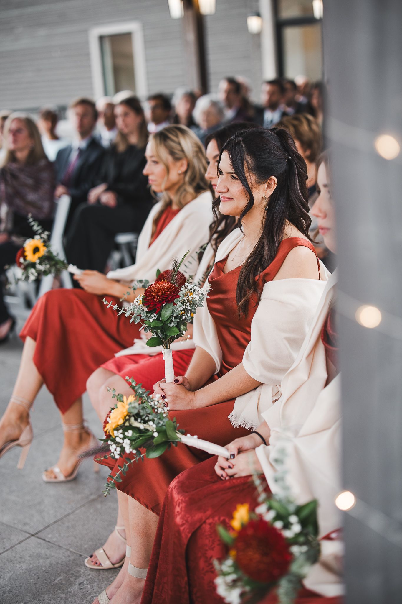 Bridesmaids in elegant red dresses sit holding bouquets of flowers at an indoor wedding ceremony.
