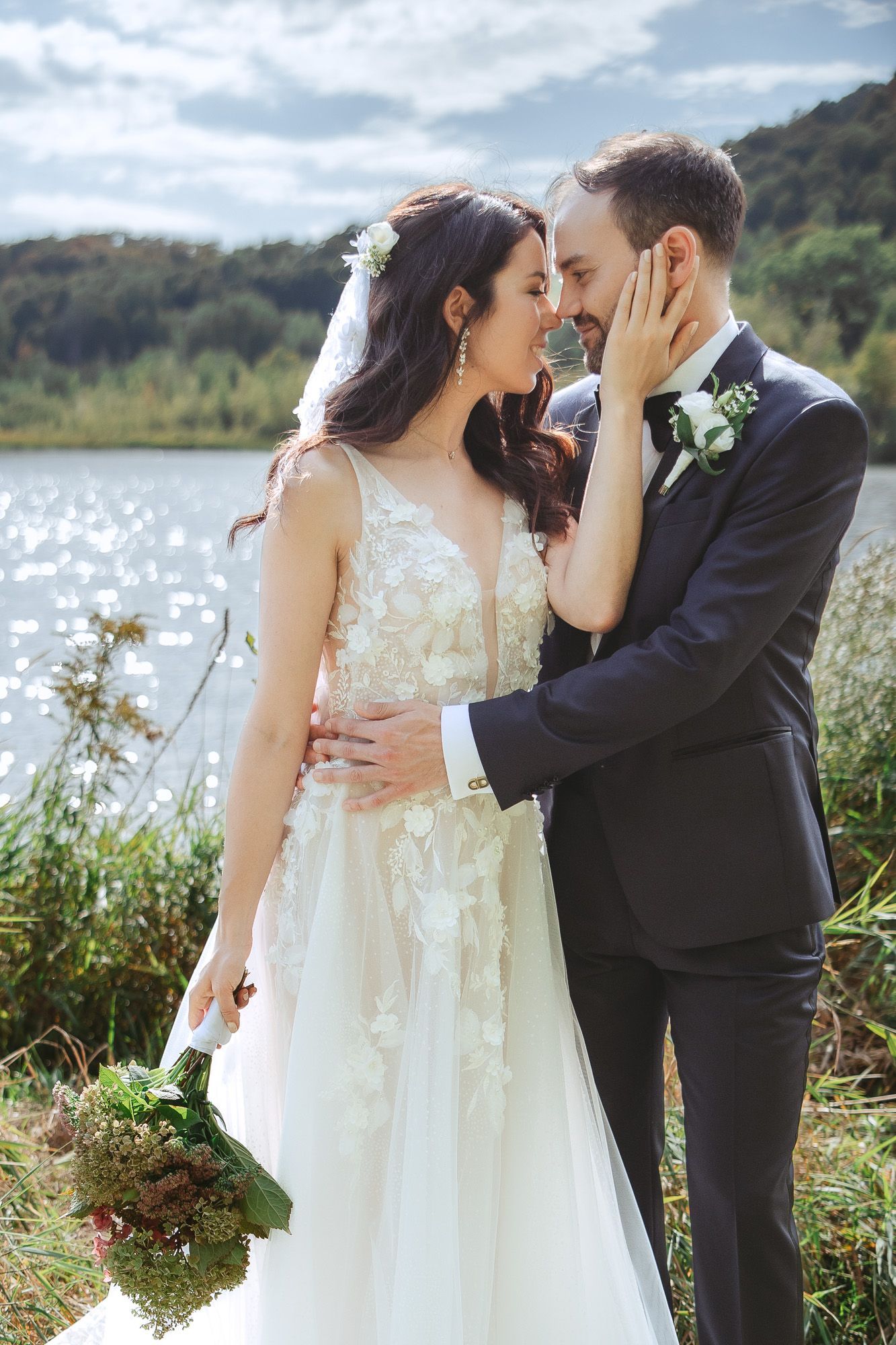 Couple de mariés s’enlaçant au bord d’un lac paisible, la mariée en robe de dentelle fluide à longue traîne, à Bromont
