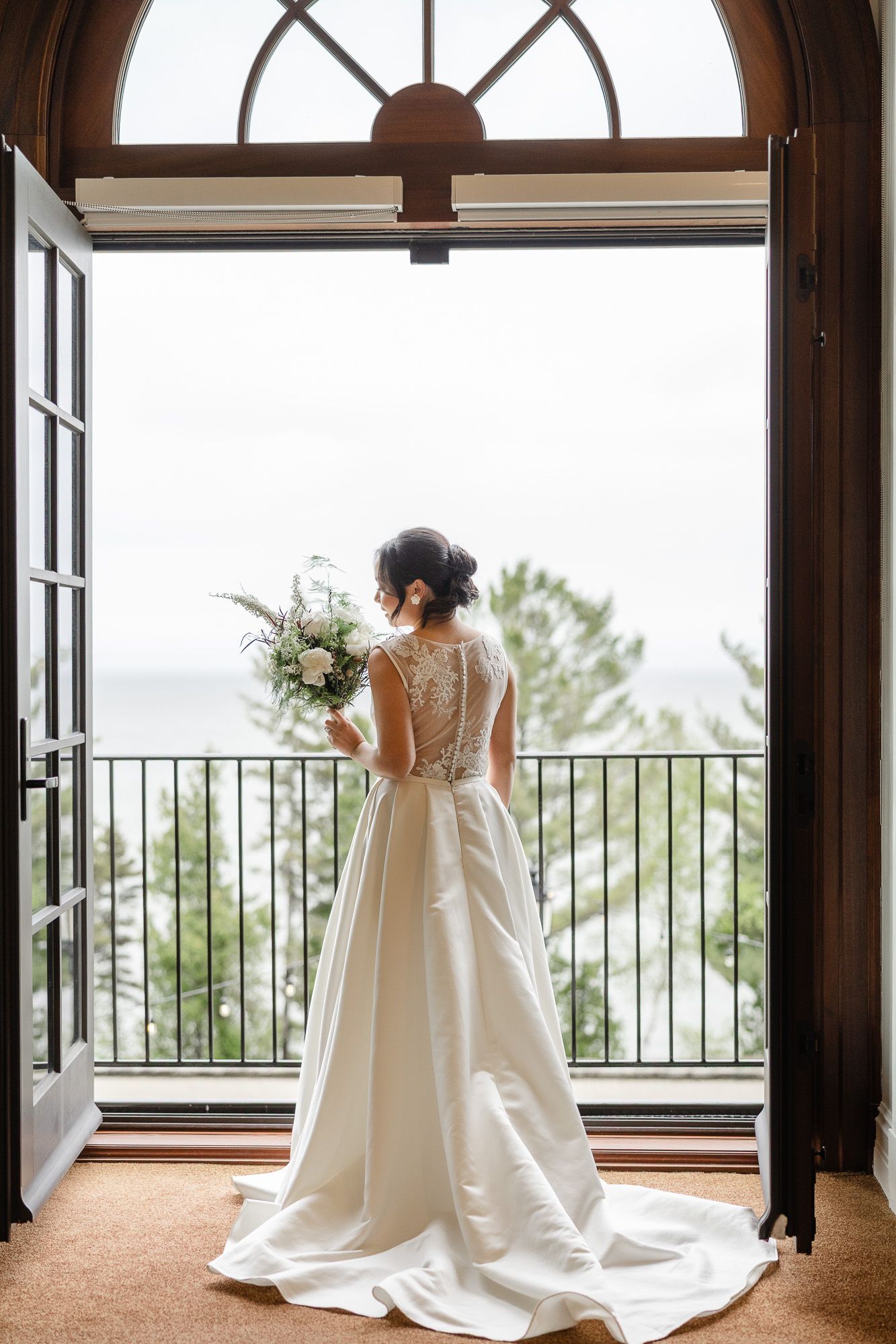 Mariée en robe blanche fluide se tenant près de grandes fenêtres avec vue sur un paysage extérieur au Manoir Richelieu