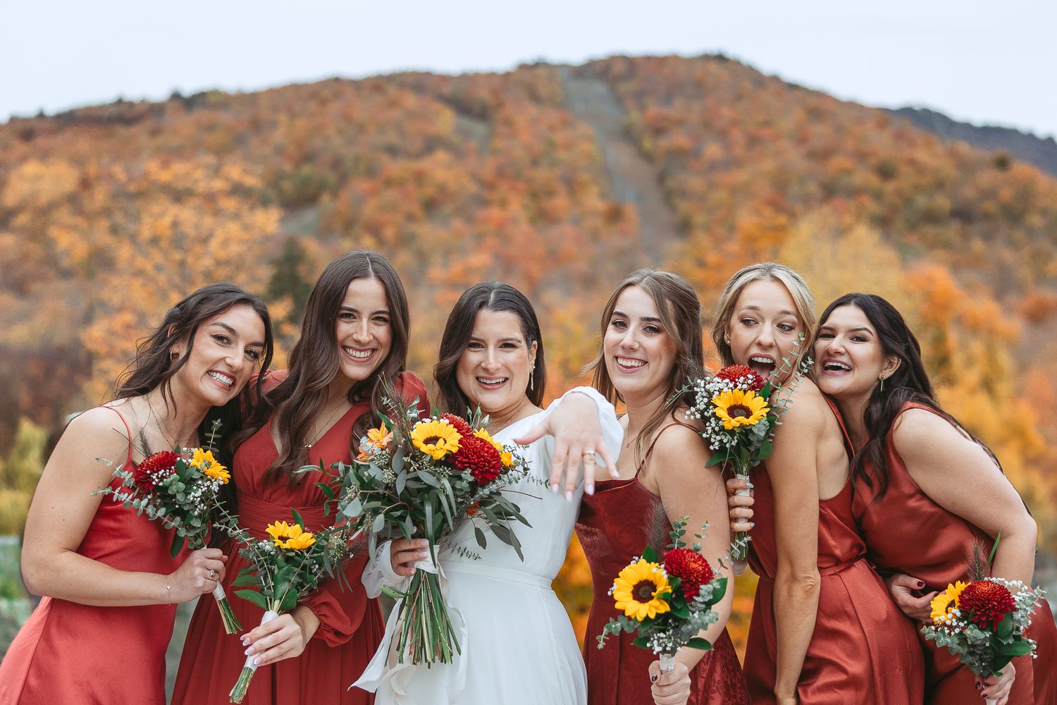 Un groupe vêtu de robes bourgogne tient des bouquets floraux colorés devant un paysage montagneux aux teintes automnales.