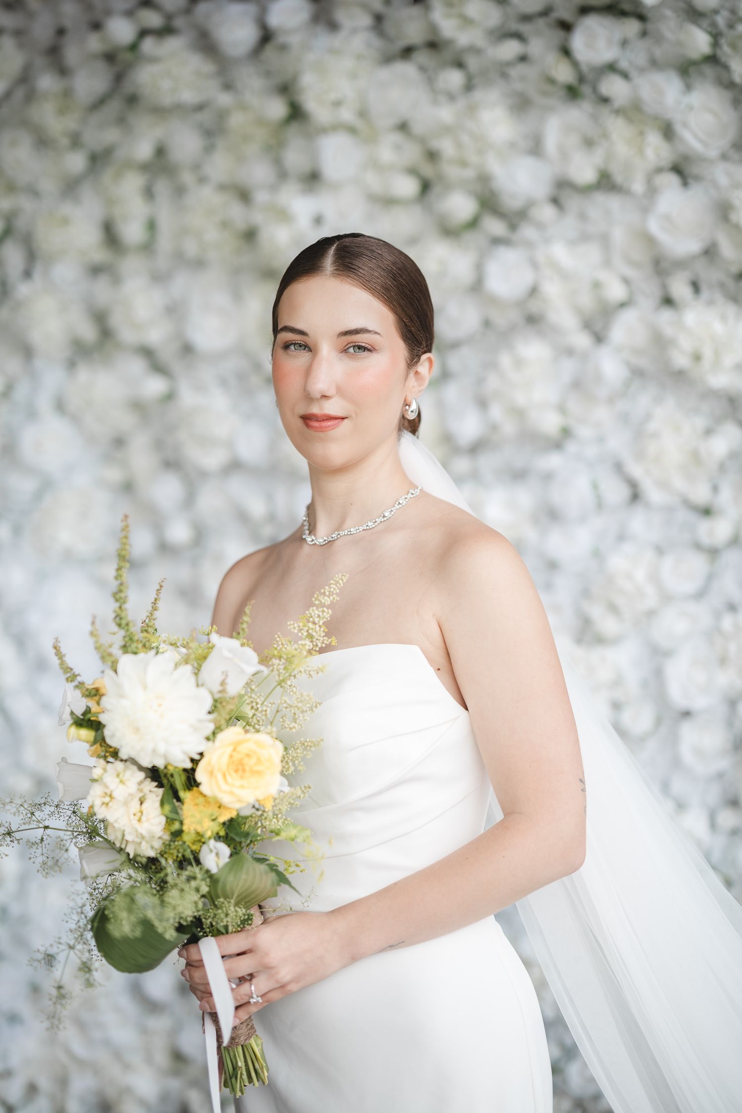 Une mariée en robe blanche bustier tient un bouquet délicat de fleurs blanches et jaunes devant un décor floral au Lac-Brome