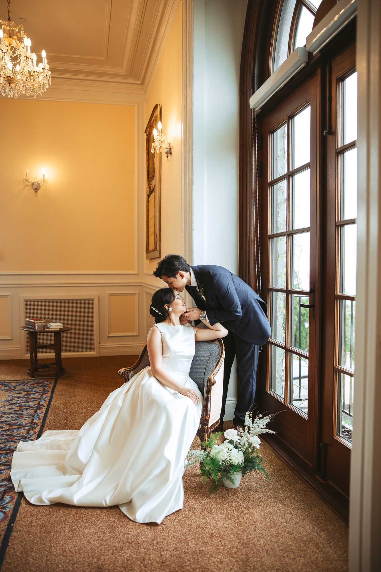 Couple de mariés partageant un moment intime dans une salle de bal élégante avec lustres en cristal et grandes fenêtres