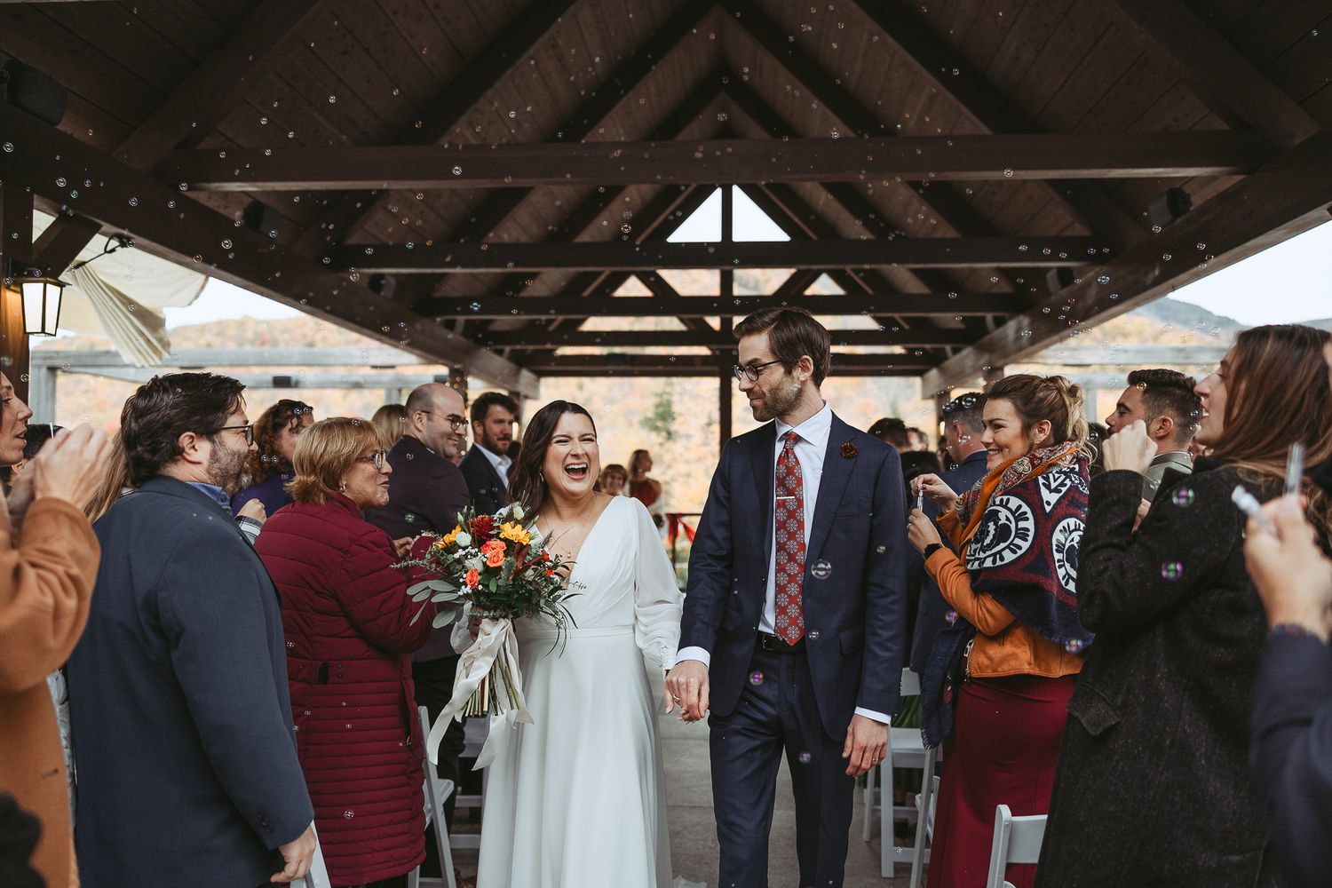 Wedding guests mingle and celebrate during an indoor reception with rustic wooden beams and string lights overhead.
