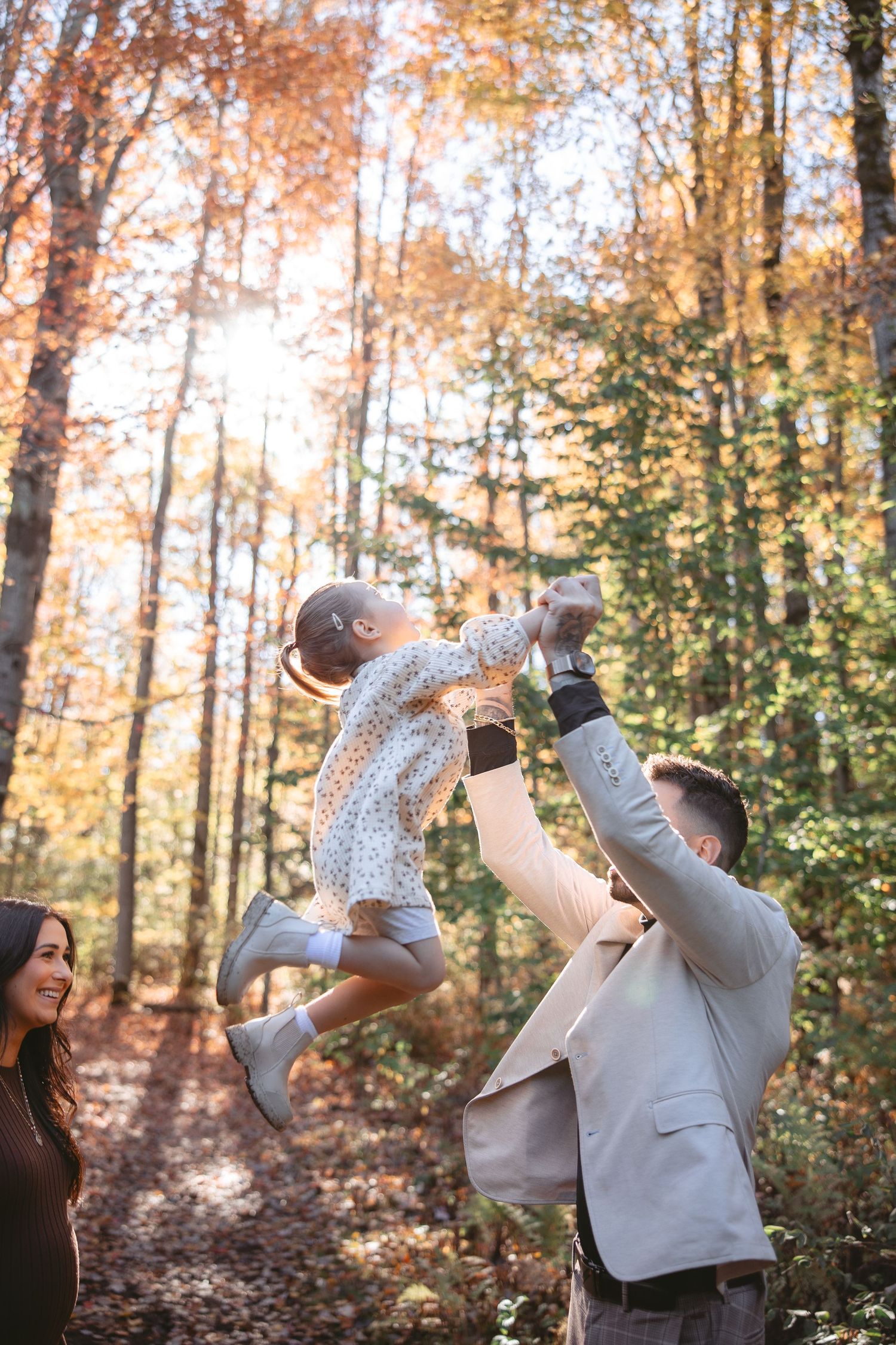Parent playfully lifts child in autumn forest with golden leaves and sunlight filtering through trees.