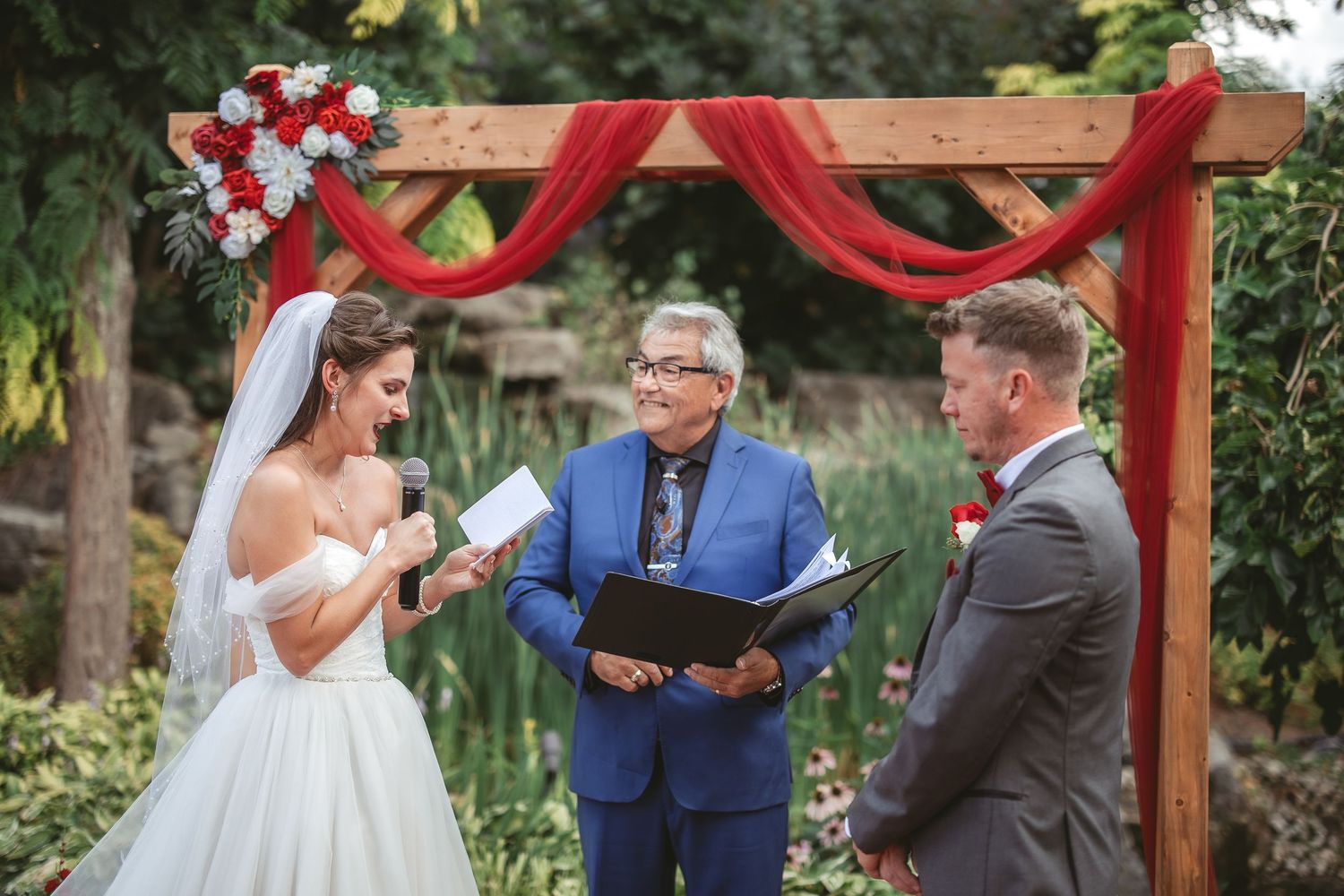 An outdoor wedding ceremony takes place under a wooden arch decorated with red fabric and floral arrangements.
