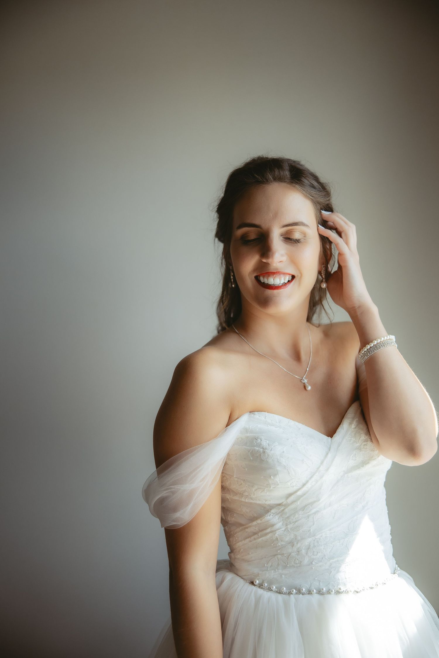 Woman in white wedding dress smiling and adjusting her hair against neutral background.