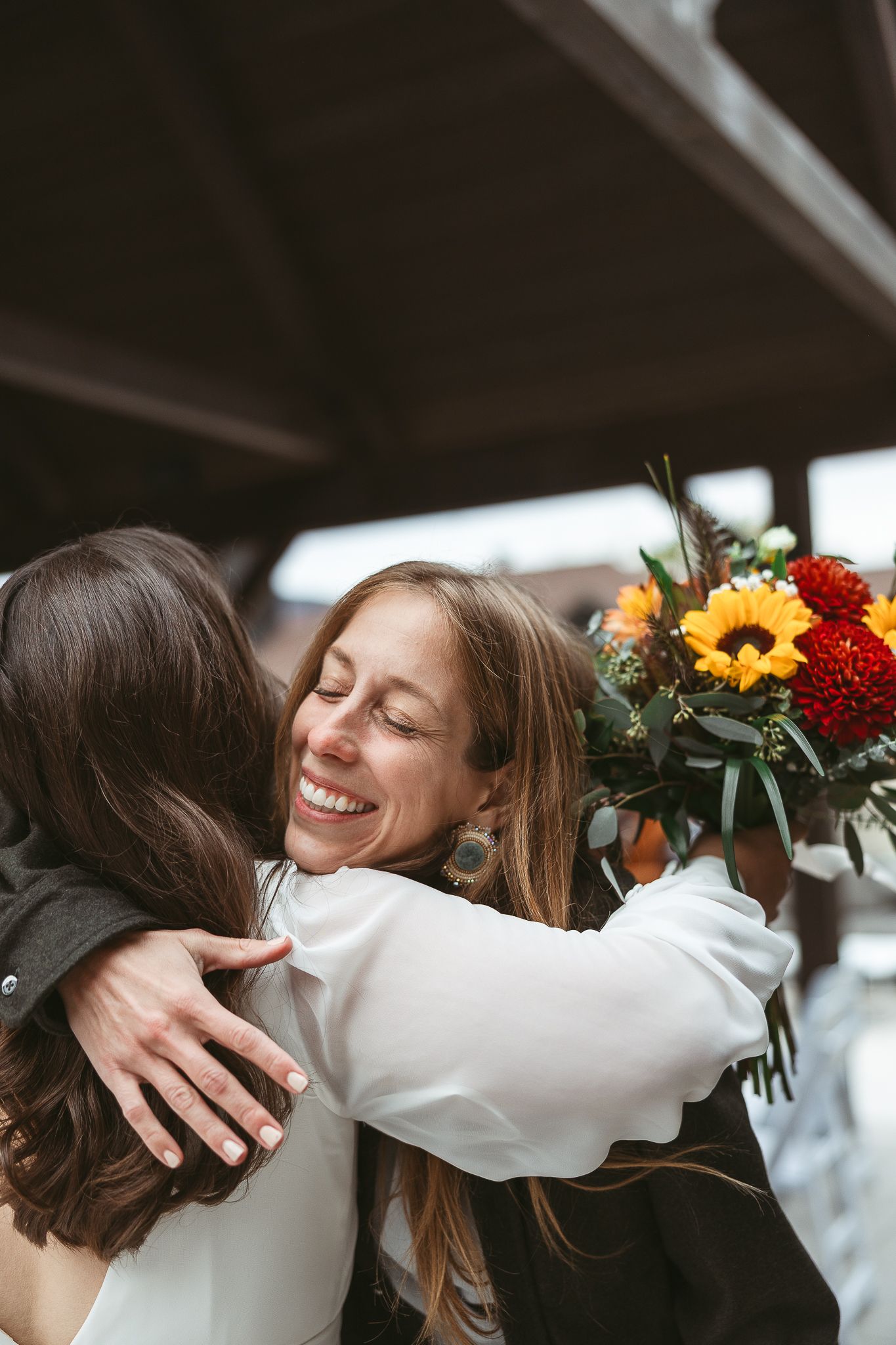 Deux personnes partagent une étreinte chaleureuse, tandis que l’une d’elles tient un bouquet vibrant de tournesols et de fleurs aux teintes