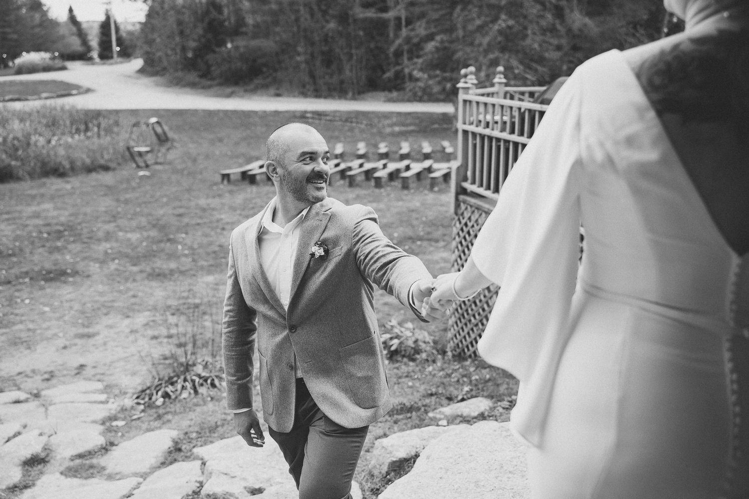 Moment joyeux en noir et blanc où un invité en veston accueille une personne vêtue de blanc sur des marches extérieures lors du mariage.