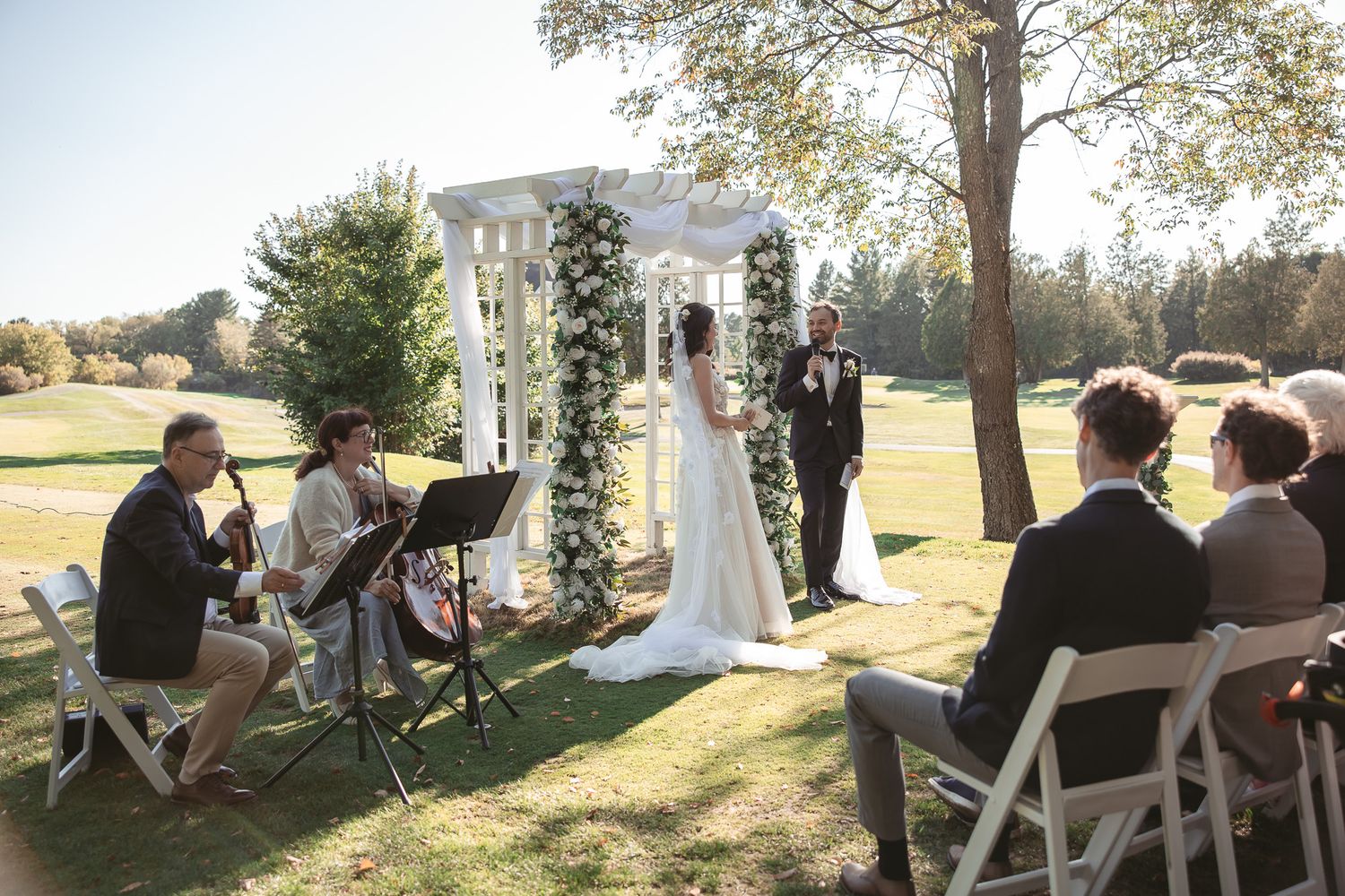 Cérémonie de mariage en plein air sous les arbres, chaises blanches installées lors d’une journée d’automne ensoleillée, en Estrie