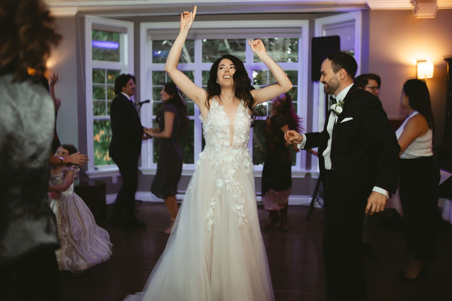 Mariée joyeuse dansant les bras levés sur une piste en bois foncé, robe de mariée à appliqués floraux