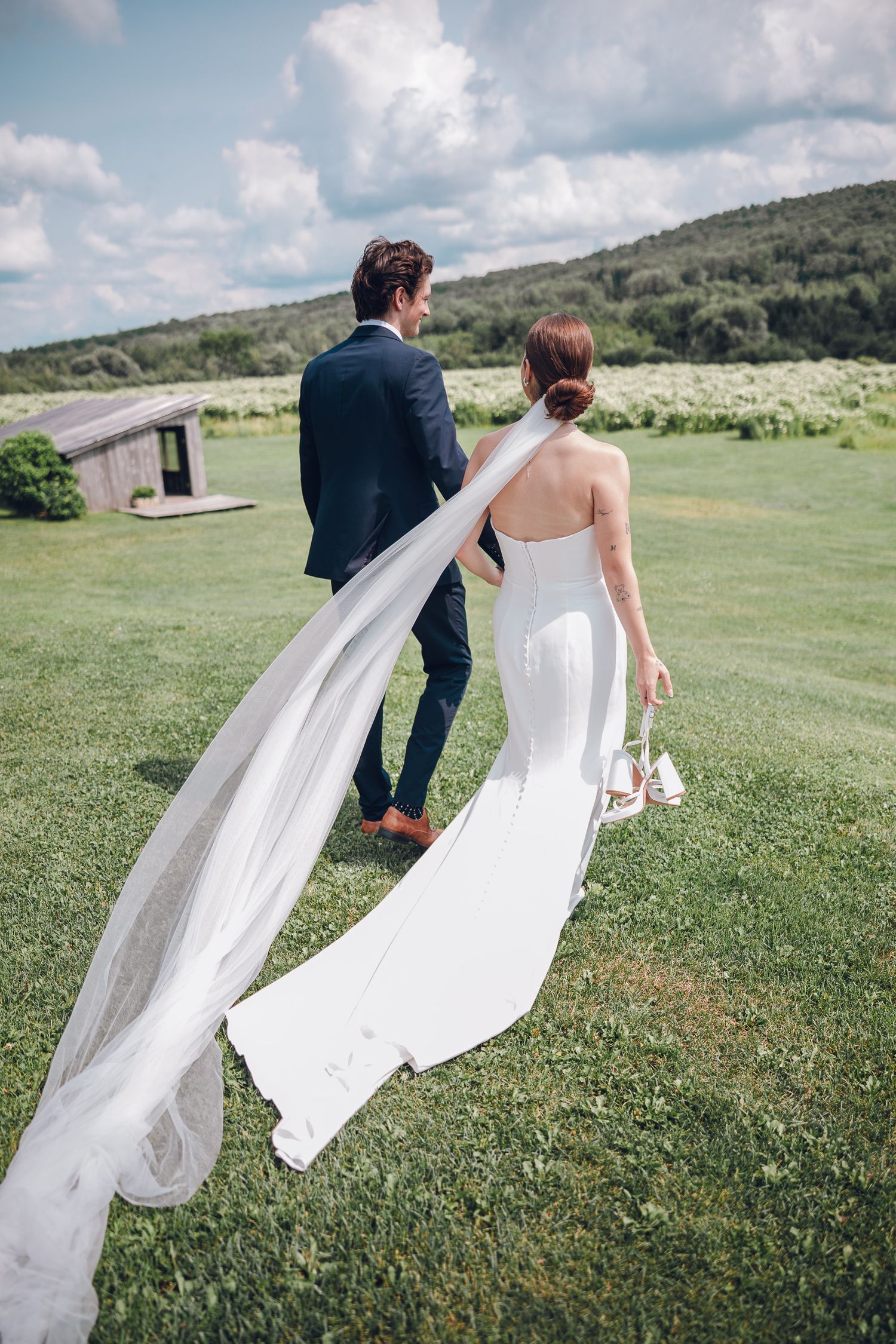 Couple de mariés qui marche vu de dos avec en arrière plan des montagnes et champ de sureaux au Lac-Brome