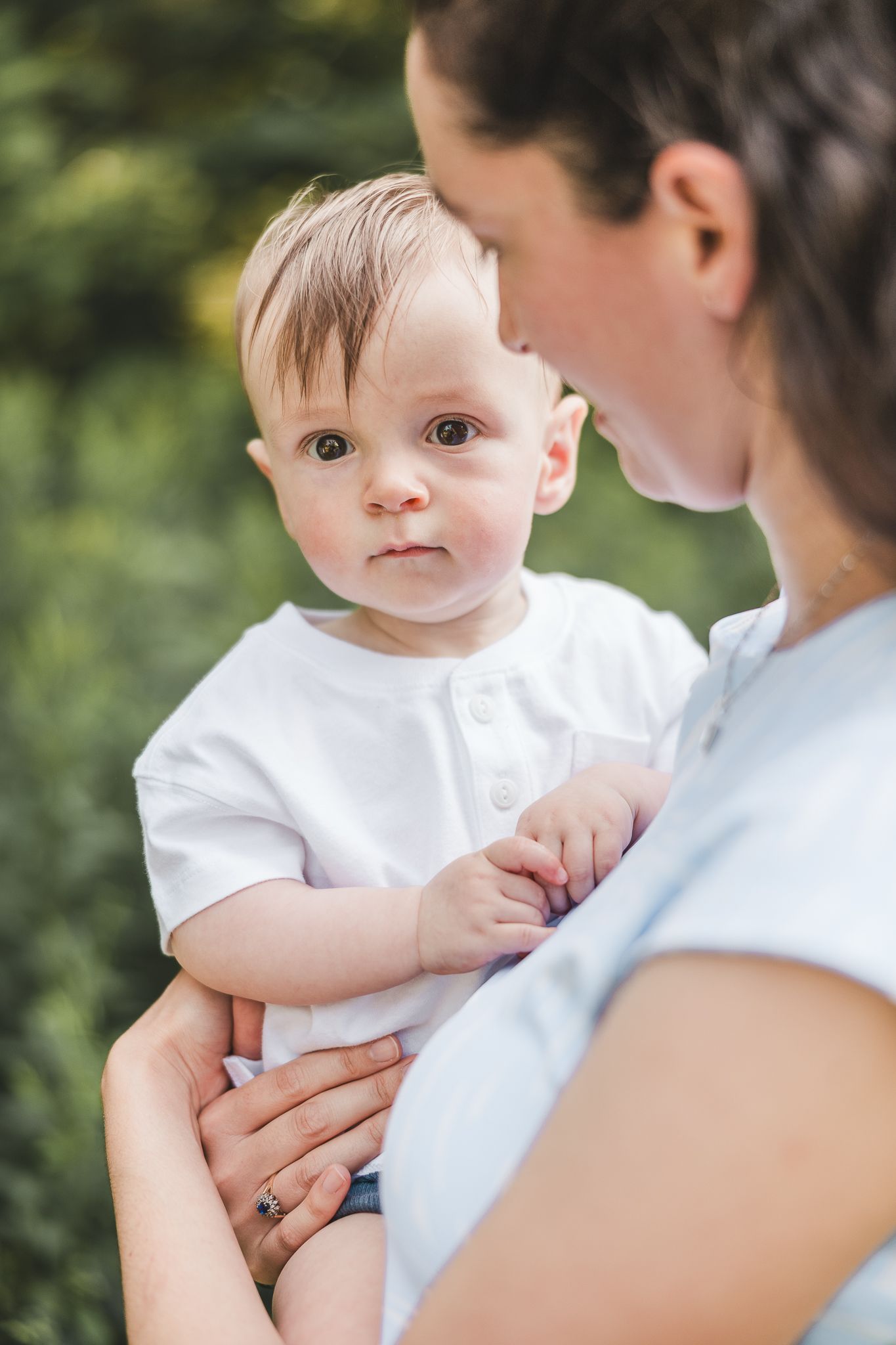Bébé tenu par parent en haut clair dans cadre extérieur verdoyant, capturé pour moments chaleureux, tendres et familiaux