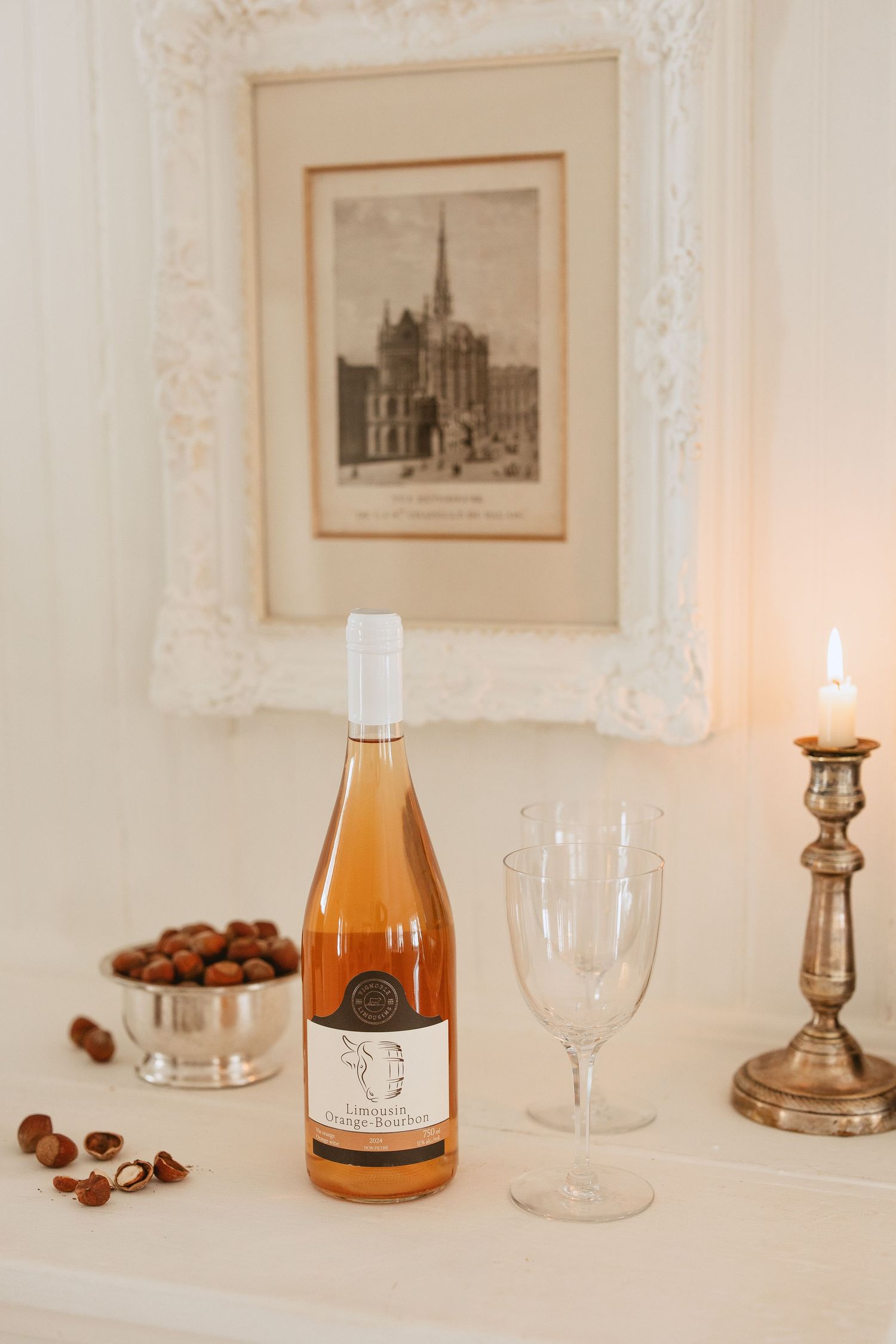 Vintage artwork frames on white wall with wine bottle, glasses, candle and hazelnuts on elegant table setting.