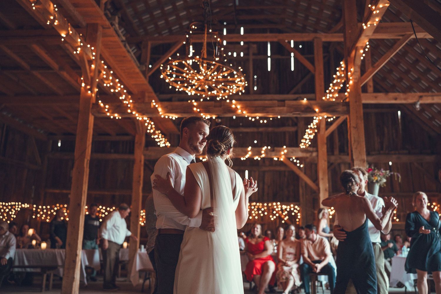 Invités dansant et célébrant un mariage sous des poutres en bois avec guirlandes lumineuses dans une grange en Estrie