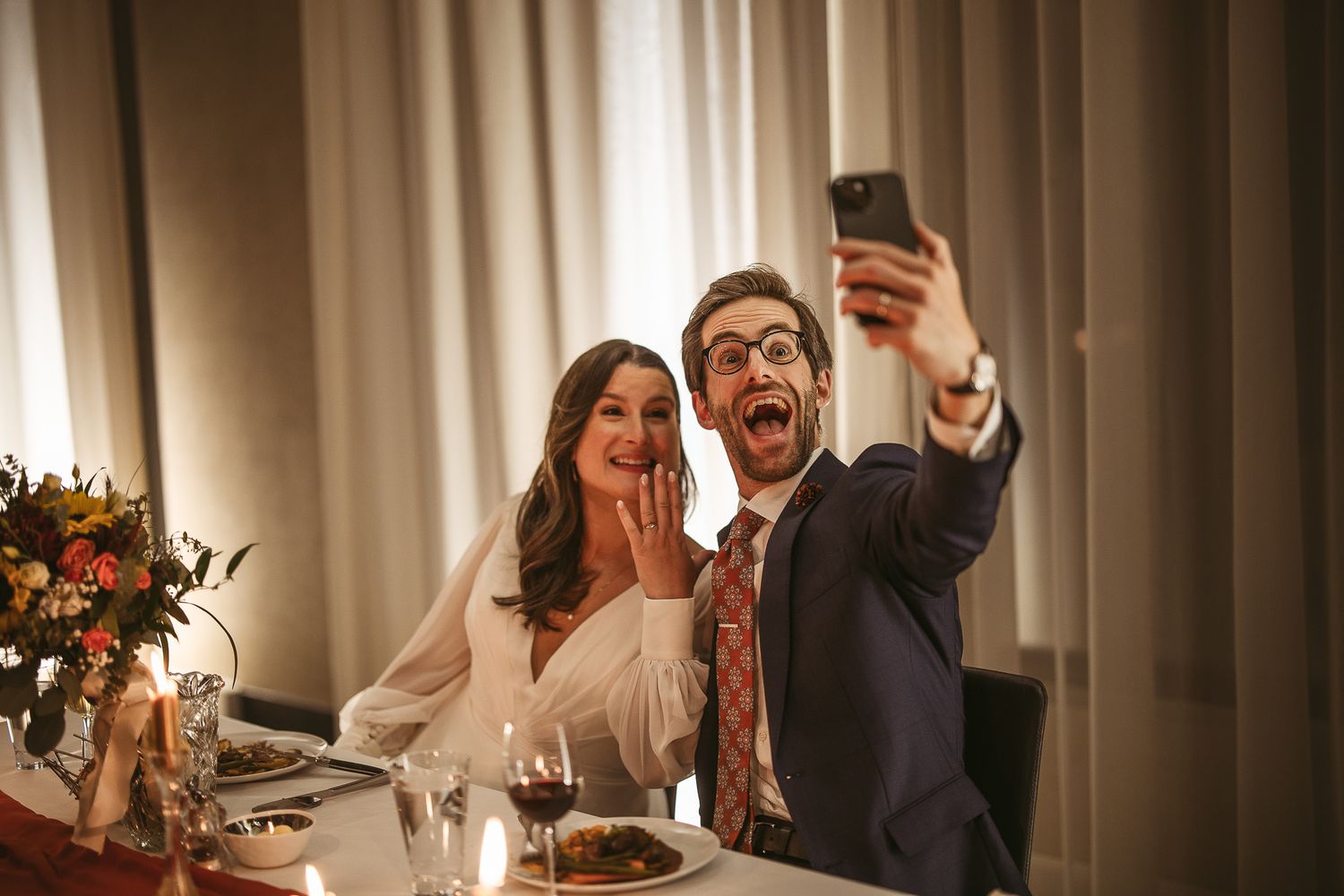 Des personnes célèbrent joyeusement lors d’un dîner, levant leurs verres et riant autour de la table en se prenant en photo.