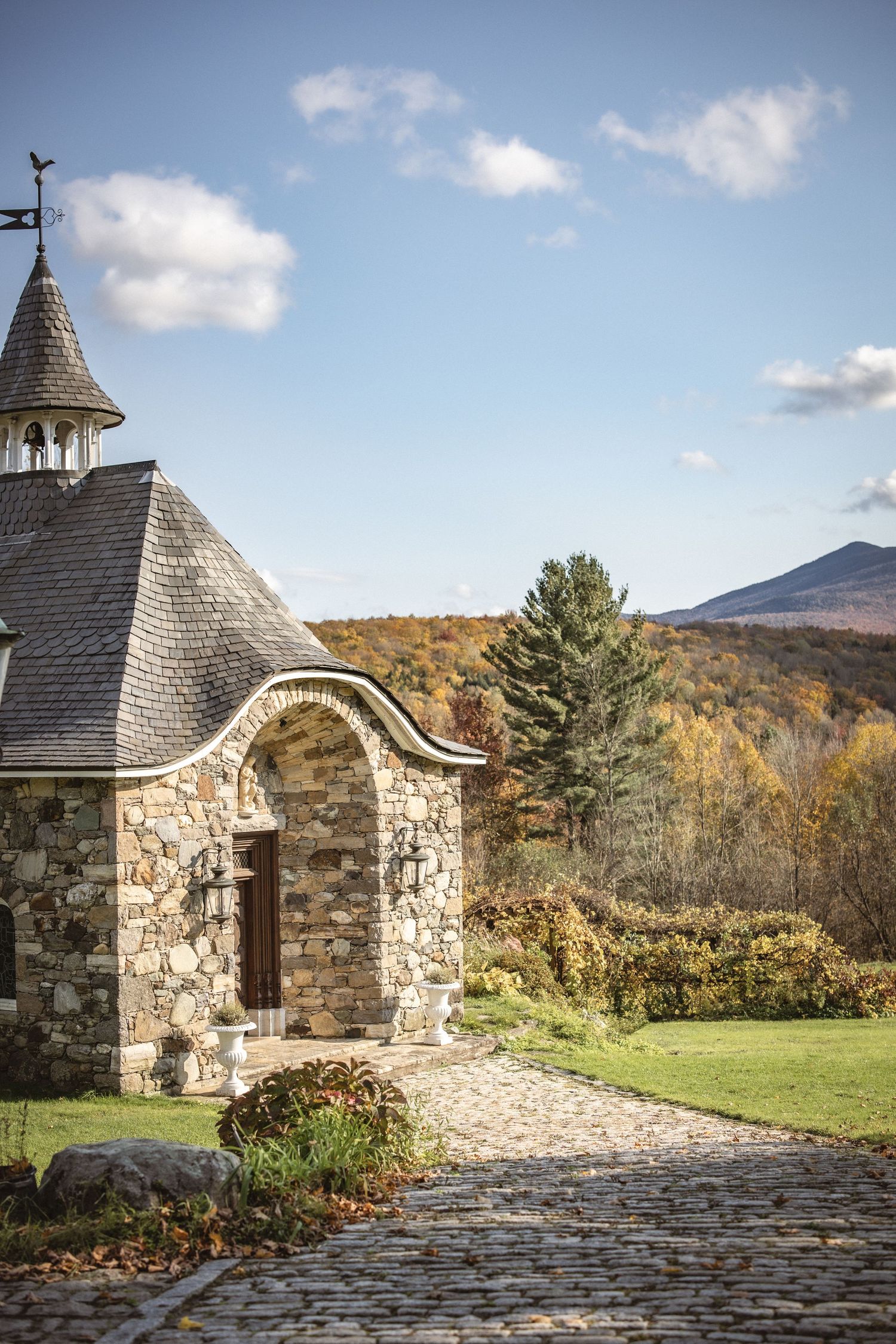 A rustic stone chapel with a spired tower sits on a hillside with autumn mountains and blue skies in the background.
