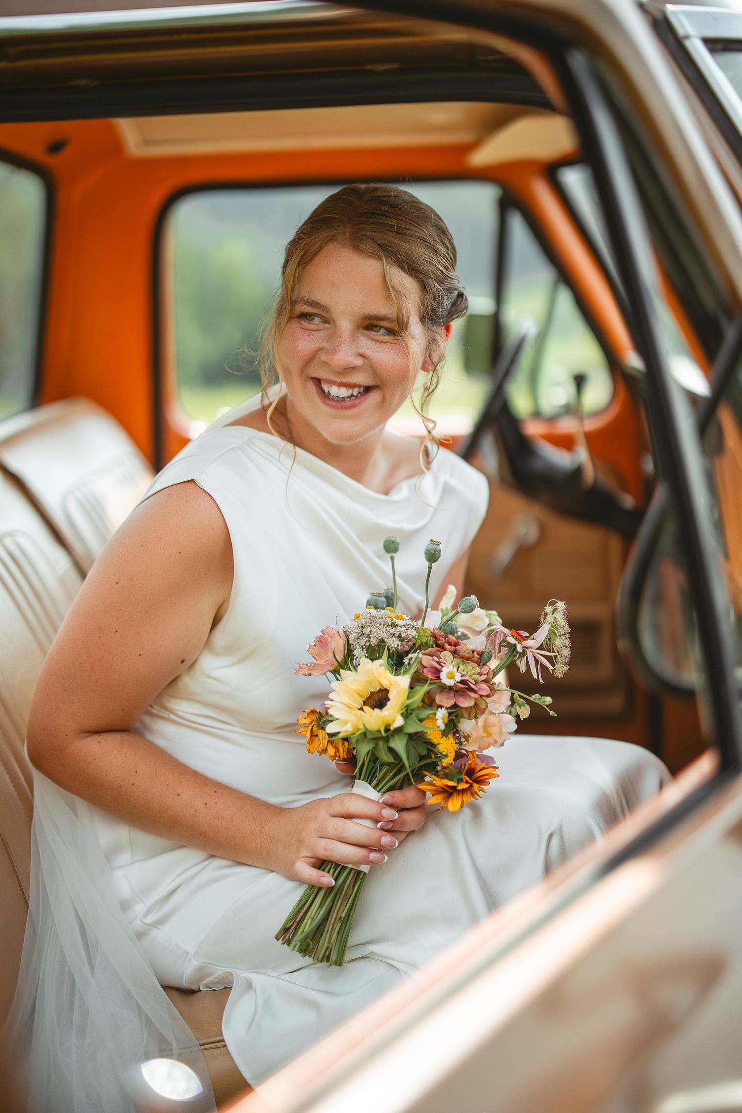 Bouquet de mariée coloré avec tournesols et fleurs variées tenu dans une voiture vintage à intérieur crème