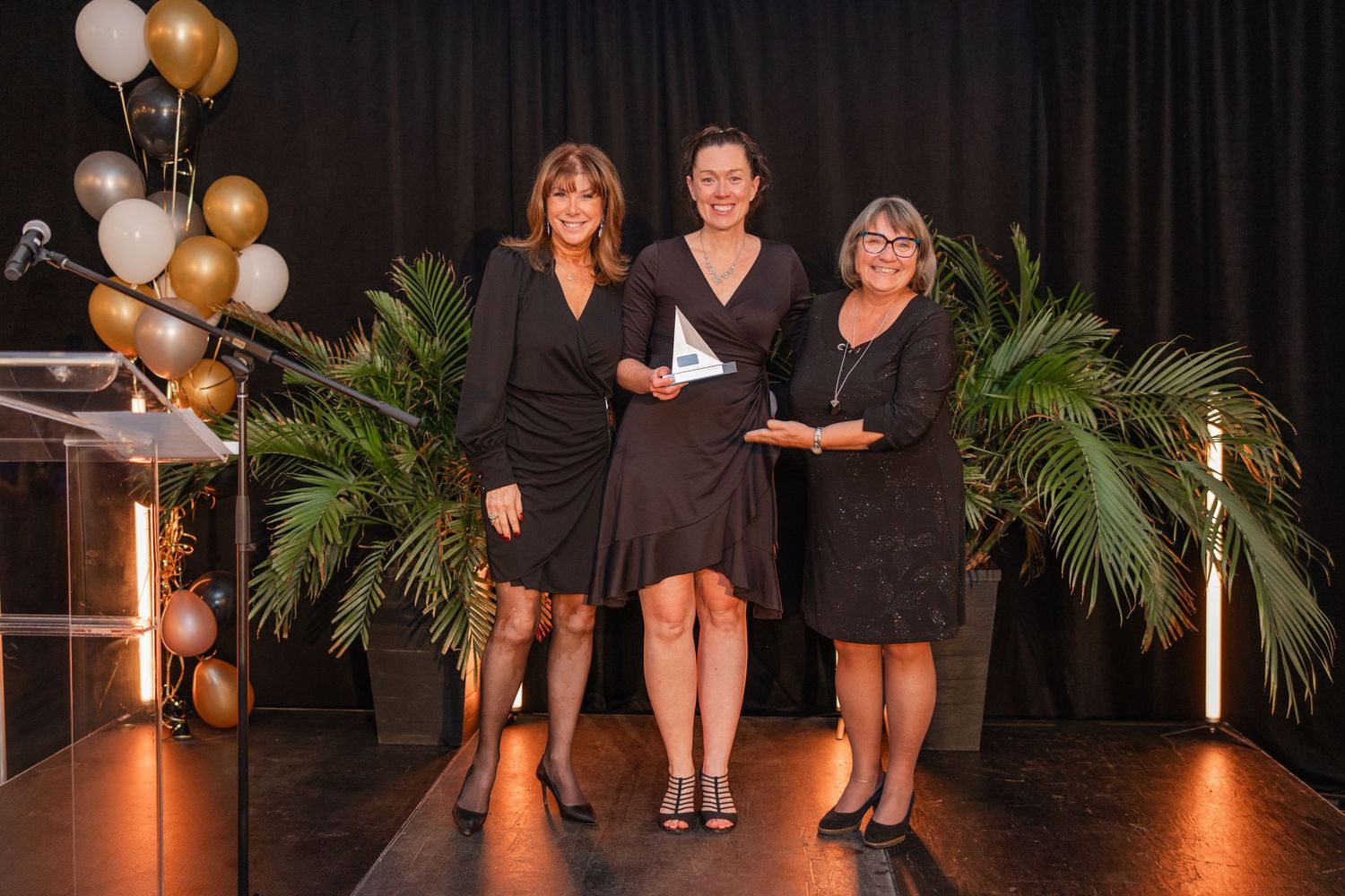 Three people in black dresses stand on stage with palm plants and gold balloons for an elegant event.