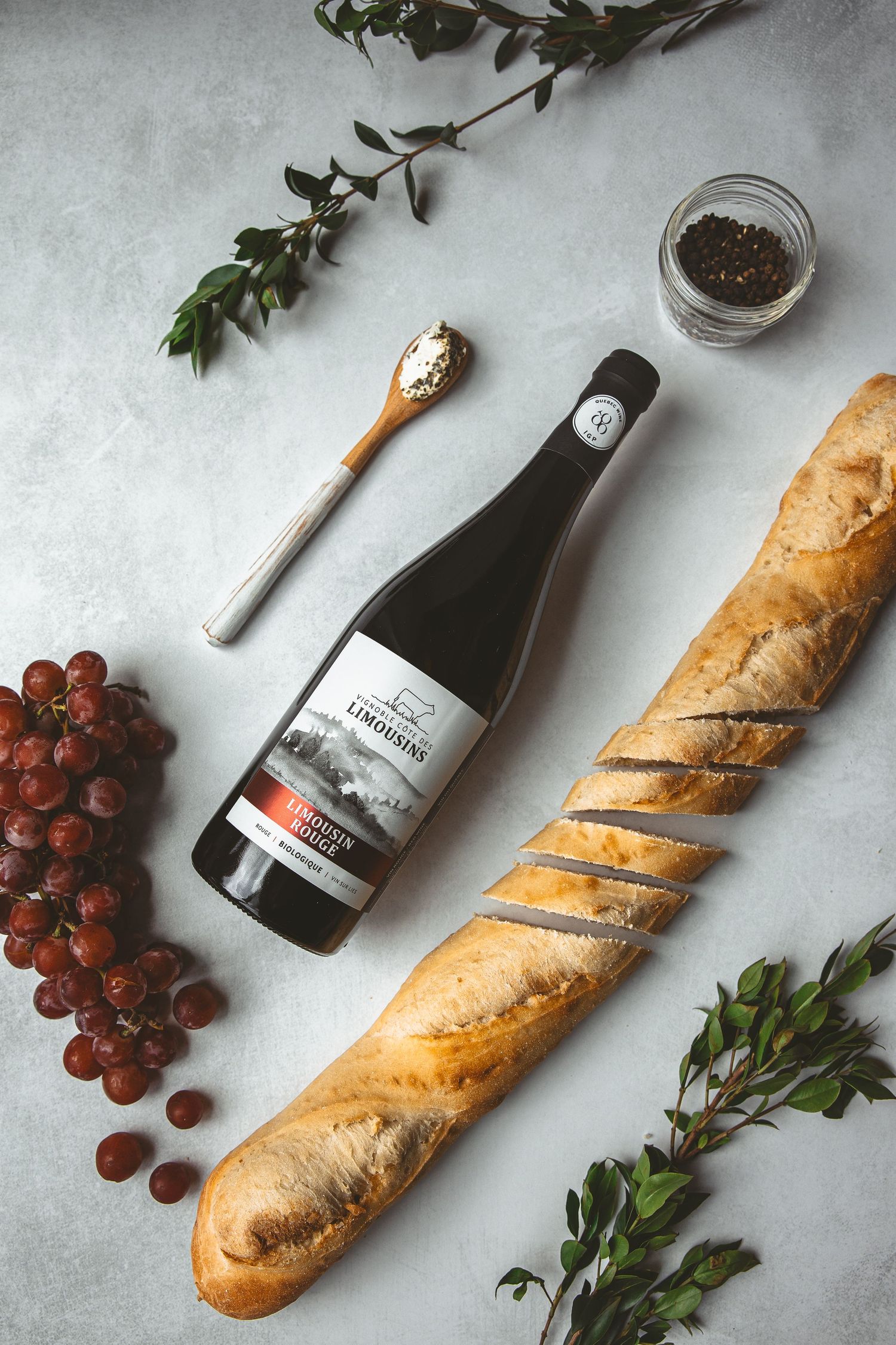 Wine bottle with fresh baguette, grapes, and herbs arranged on a gray surface.