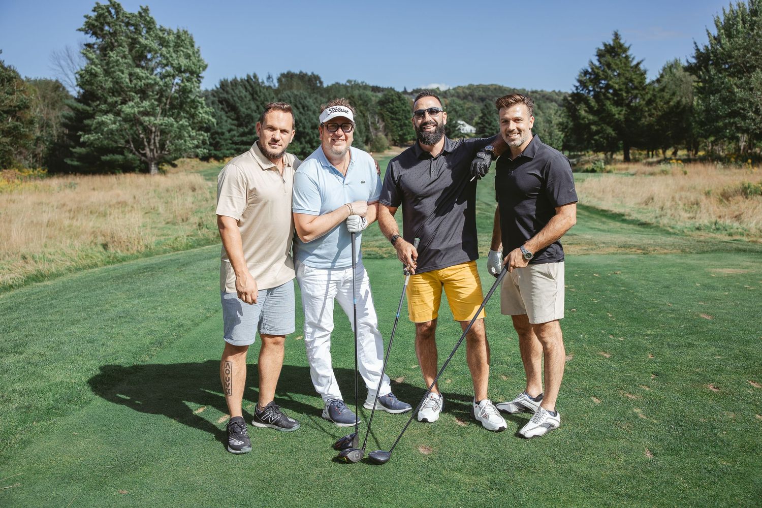 Four golfers pose together with clubs on a sunny golf course with trees and rolling hills in the background.