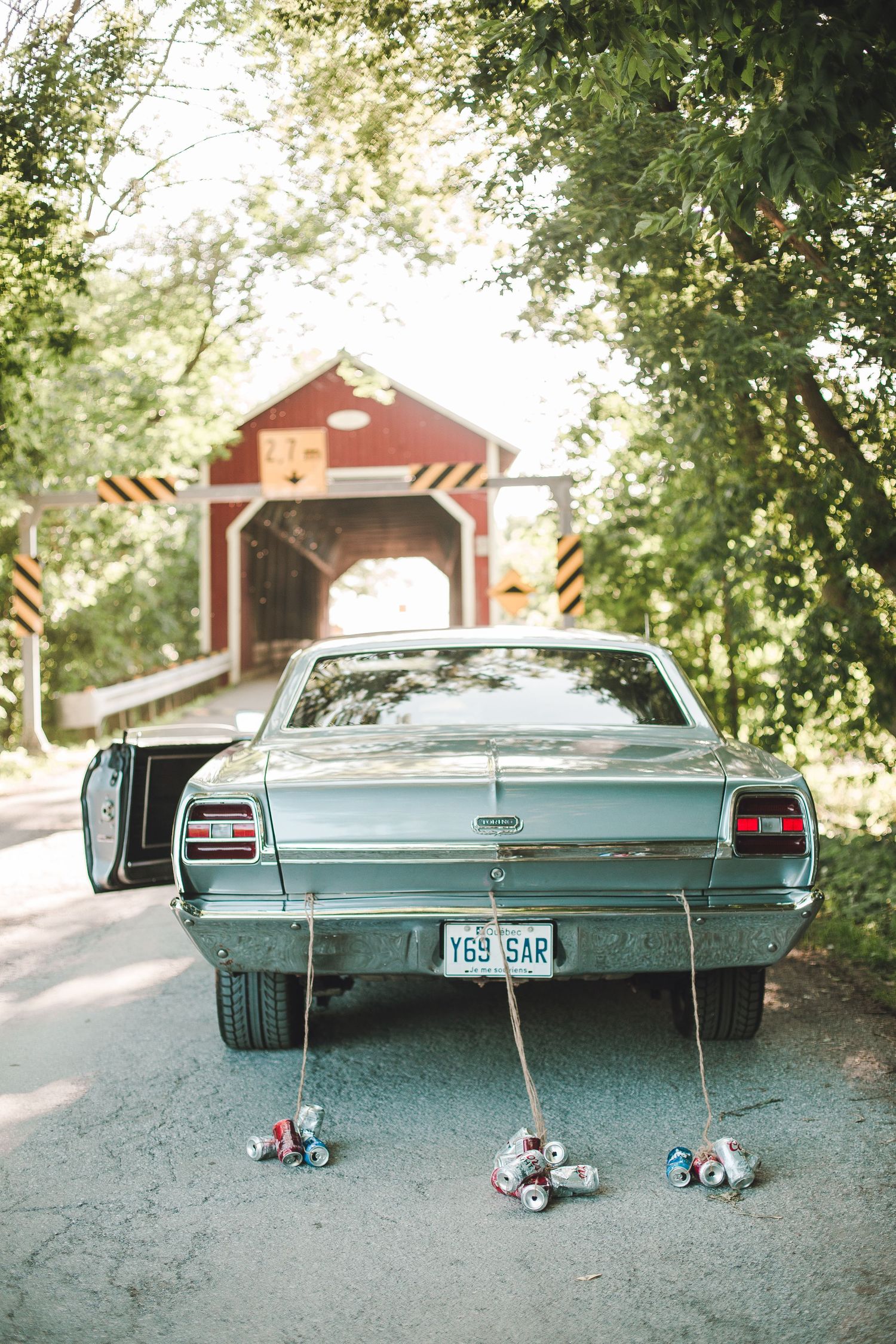 Vintage teal car with tin cans tied to bumper parked in front of red covered bridge on dirt road.