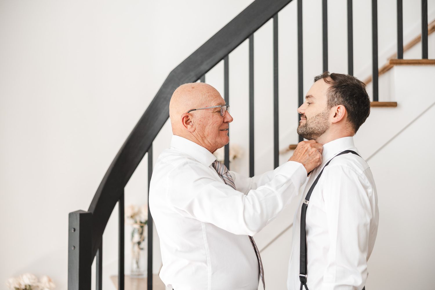 Le père et le marié partageant un moment complice en ajustant une tenue élégante près d’un escalier, mariage en Estrie