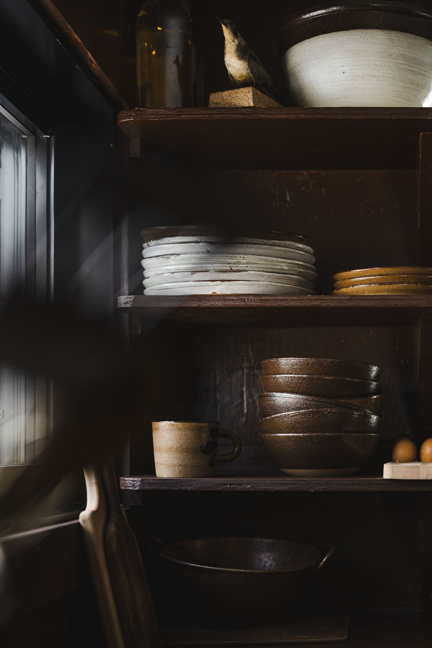 Dark wooden shelves display a collection of ceramic plates and bowls in a moody, rustic kitchen setting.