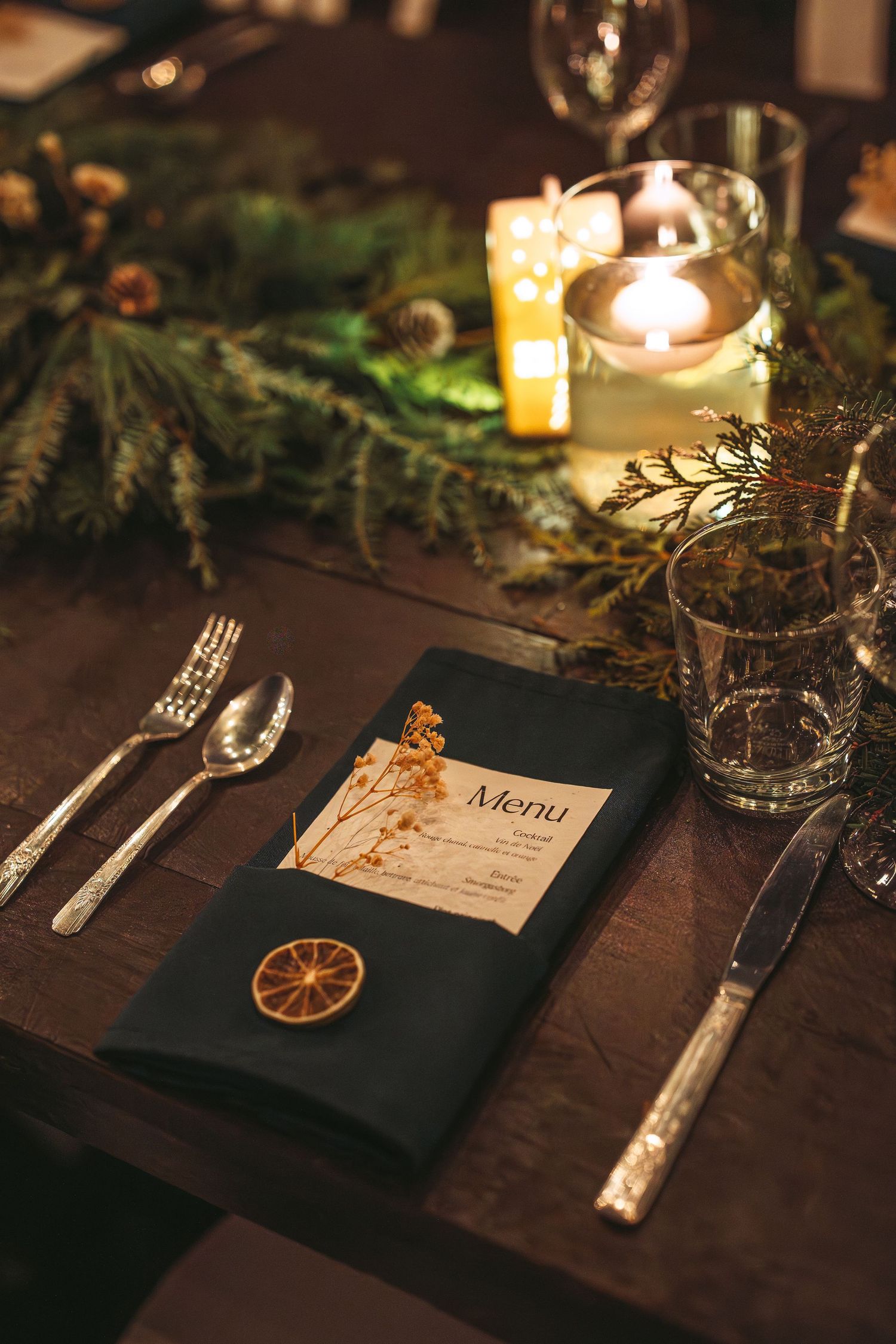 Elegant table settings with candlelight, dried citrus slices, and menu cards on a dark wooden surface at a formal dinner.