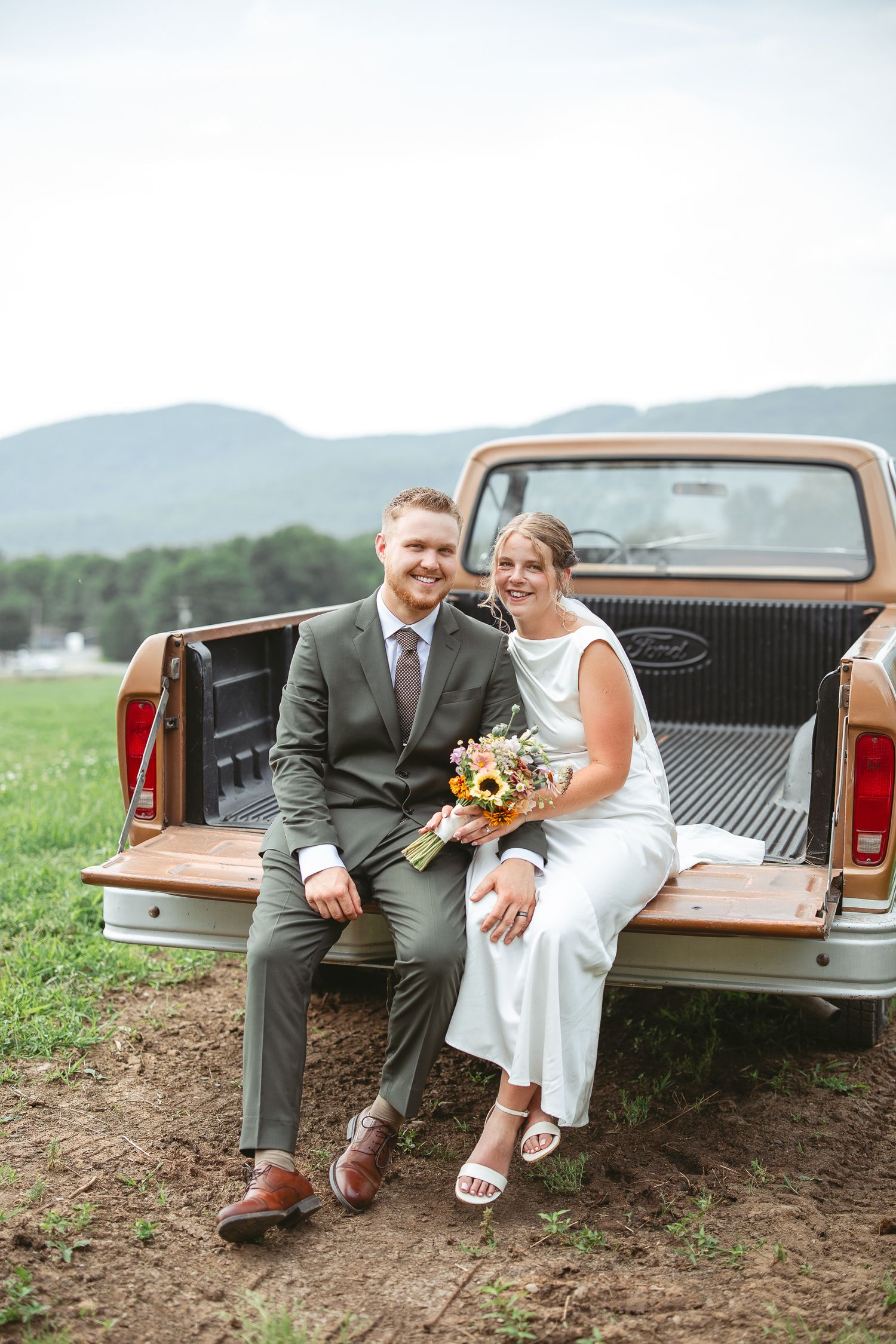 Jeunes mariés assis sur le hayon d’un pickup vintage dans un champ de montagne, avec bouquet de mariée