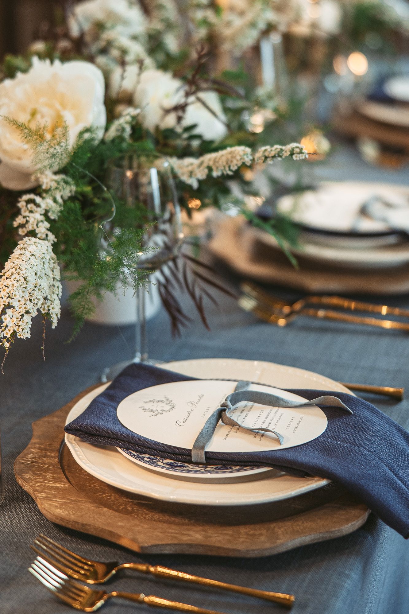 Mise en place de table élégante avec sous-assiettes en bois, serviettes bleu marine et centre de table floral blanc sur nappe grise