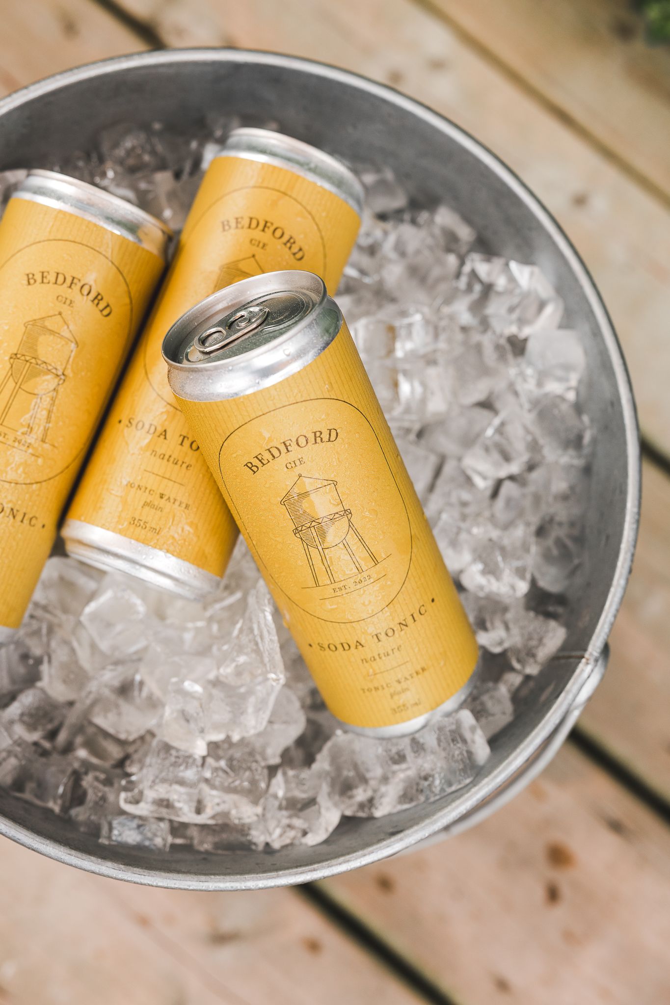 Chilled yellow beverage cans nestled in ice cubes in a metal bucket on wooden surface.