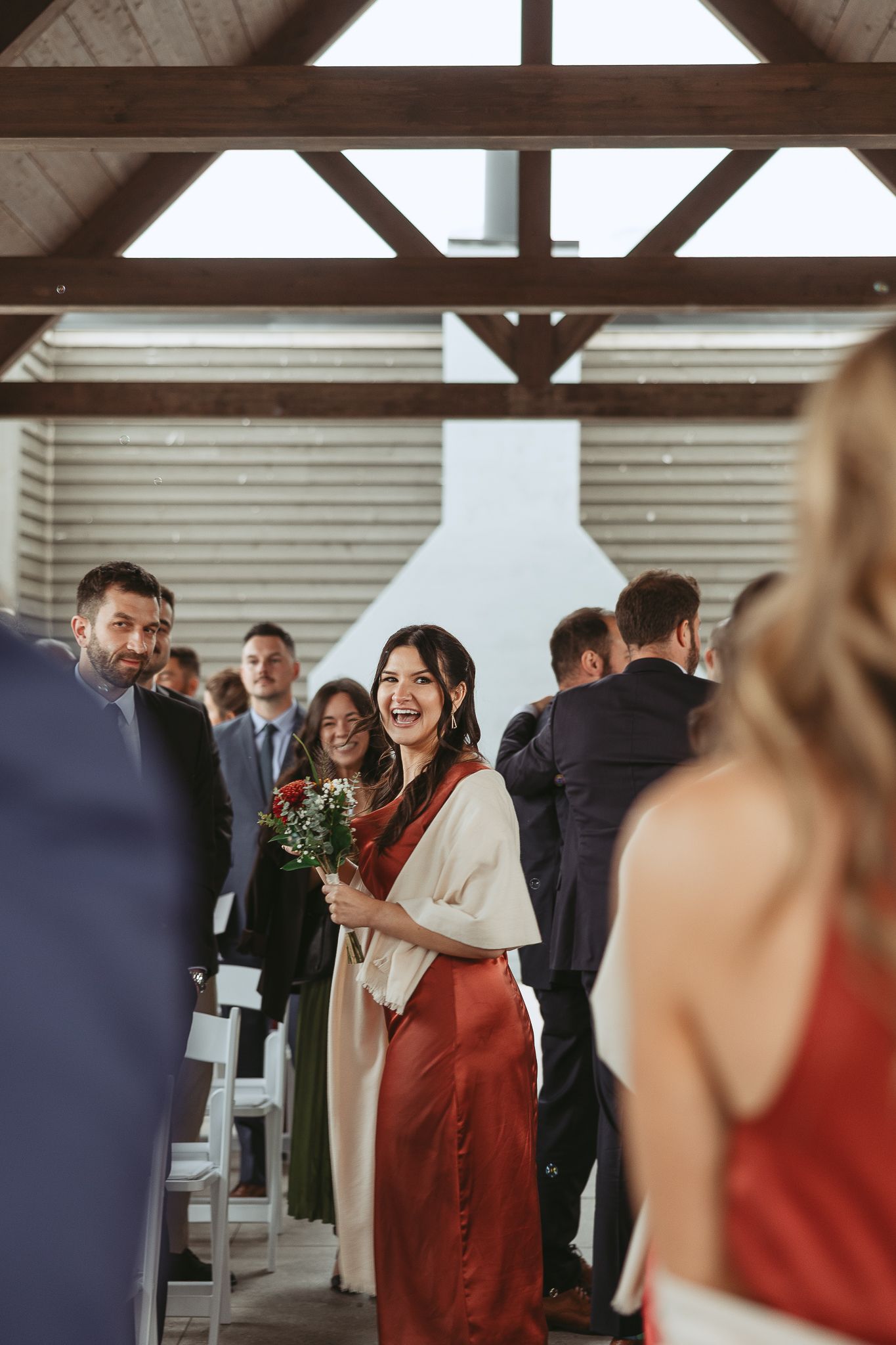 Des invités de mariage échangent et s’enlacent lors d’une réception dans un lieu intérieur rustique aux poutres de bois apparentes.