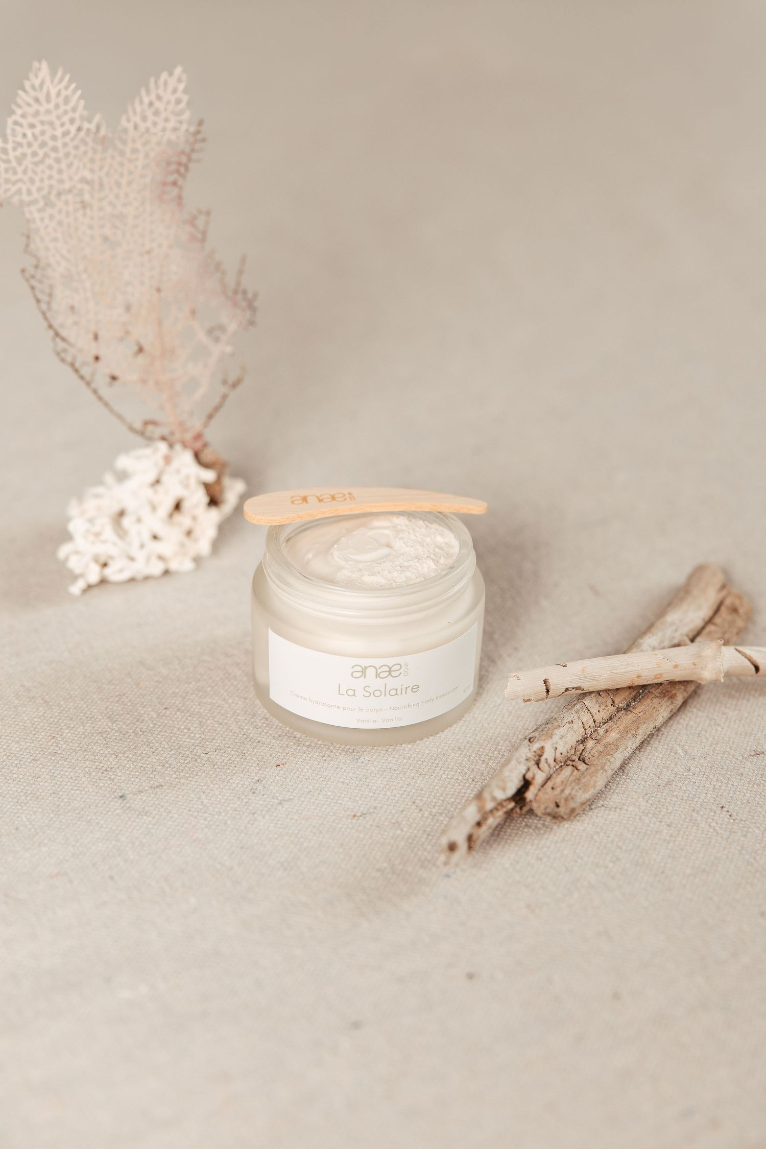 A minimalist beauty product flatlay with a white cream jar, dried coral, and driftwood on a beige textured surface.