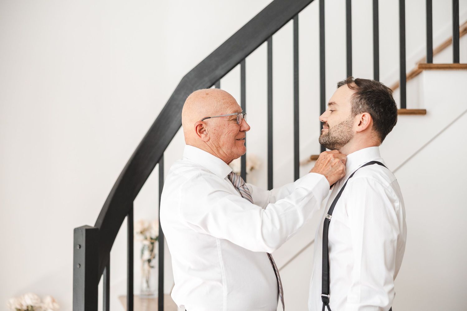 Two people stand by a staircase as one adjusts the other's bowtie in preparation for a formal event.