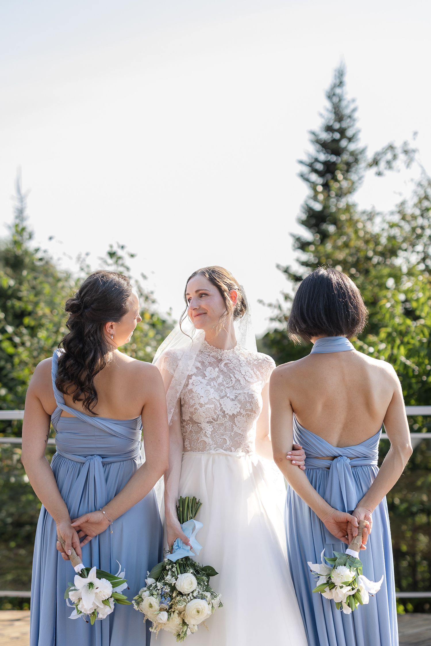 Demoiselles d’honneur en robes bleu pâle tenant des bouquets blancs, aux côtés de la mariée en robe blanche lors d’une cérémonie extérieure.