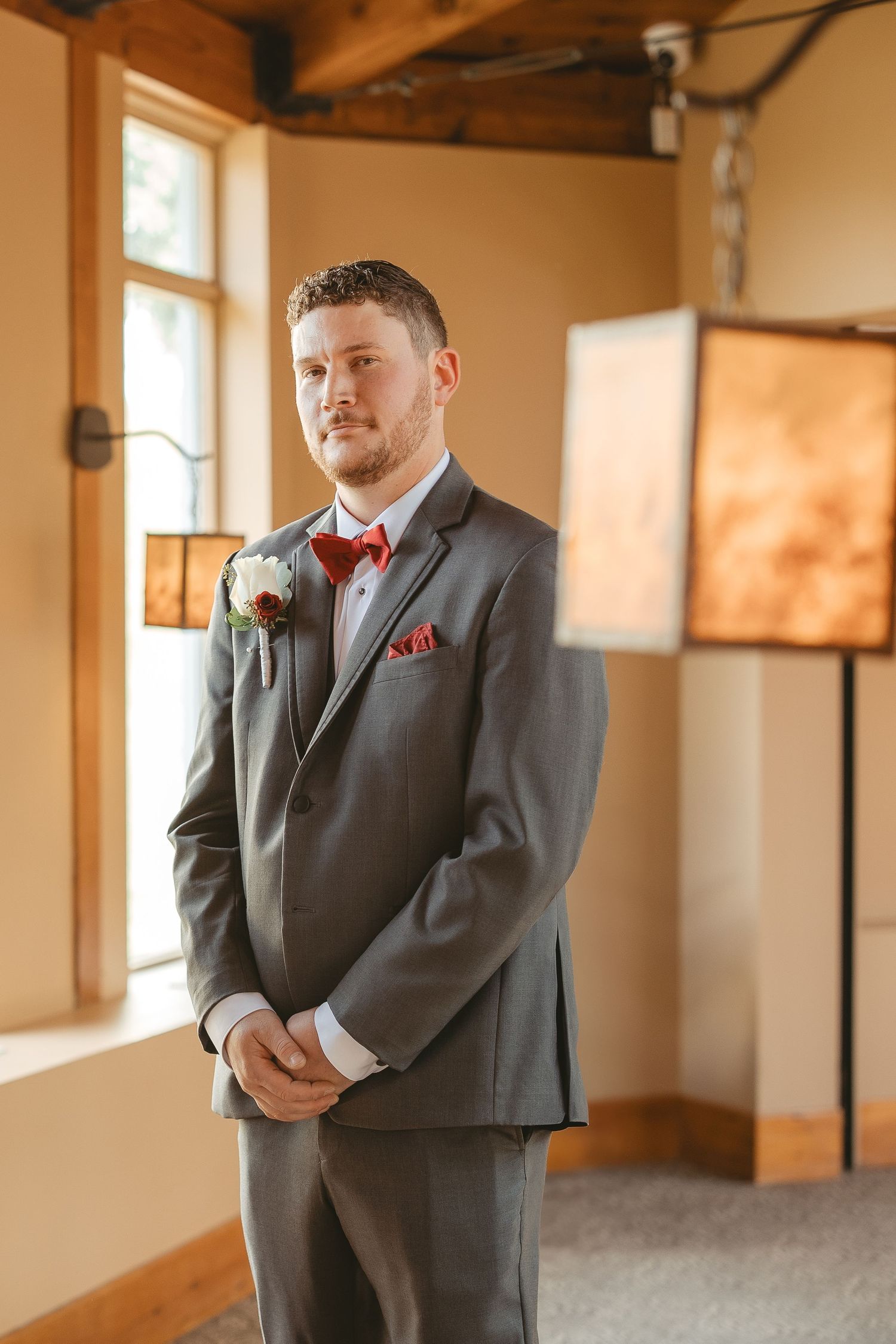 Groom in charcoal gray suit with burgundy bow tie and boutonniere poses in warm-lit indoor venue.