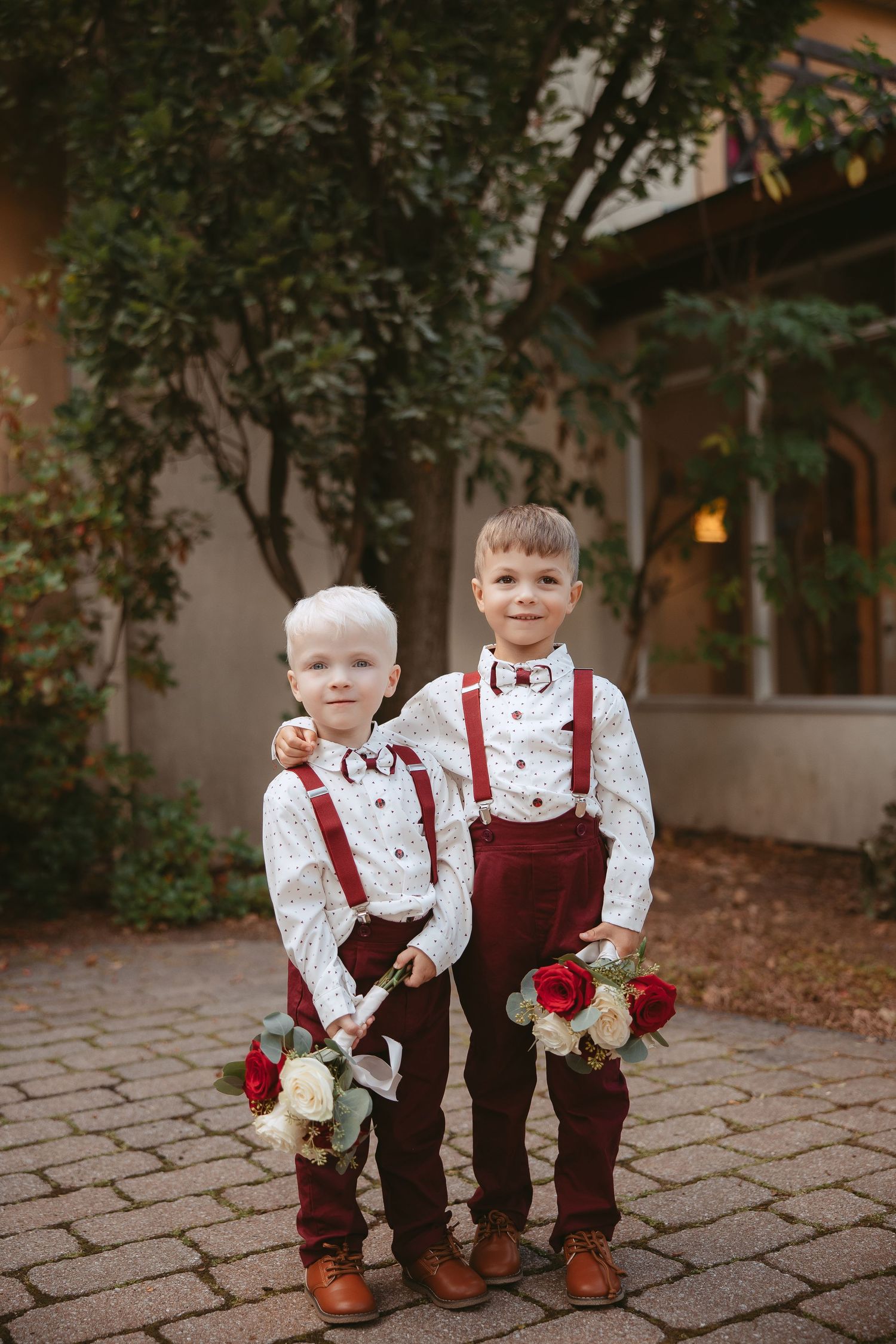 Two young ring bearers in burgundy suspenders and bow ties hold flowers on stone pathway.