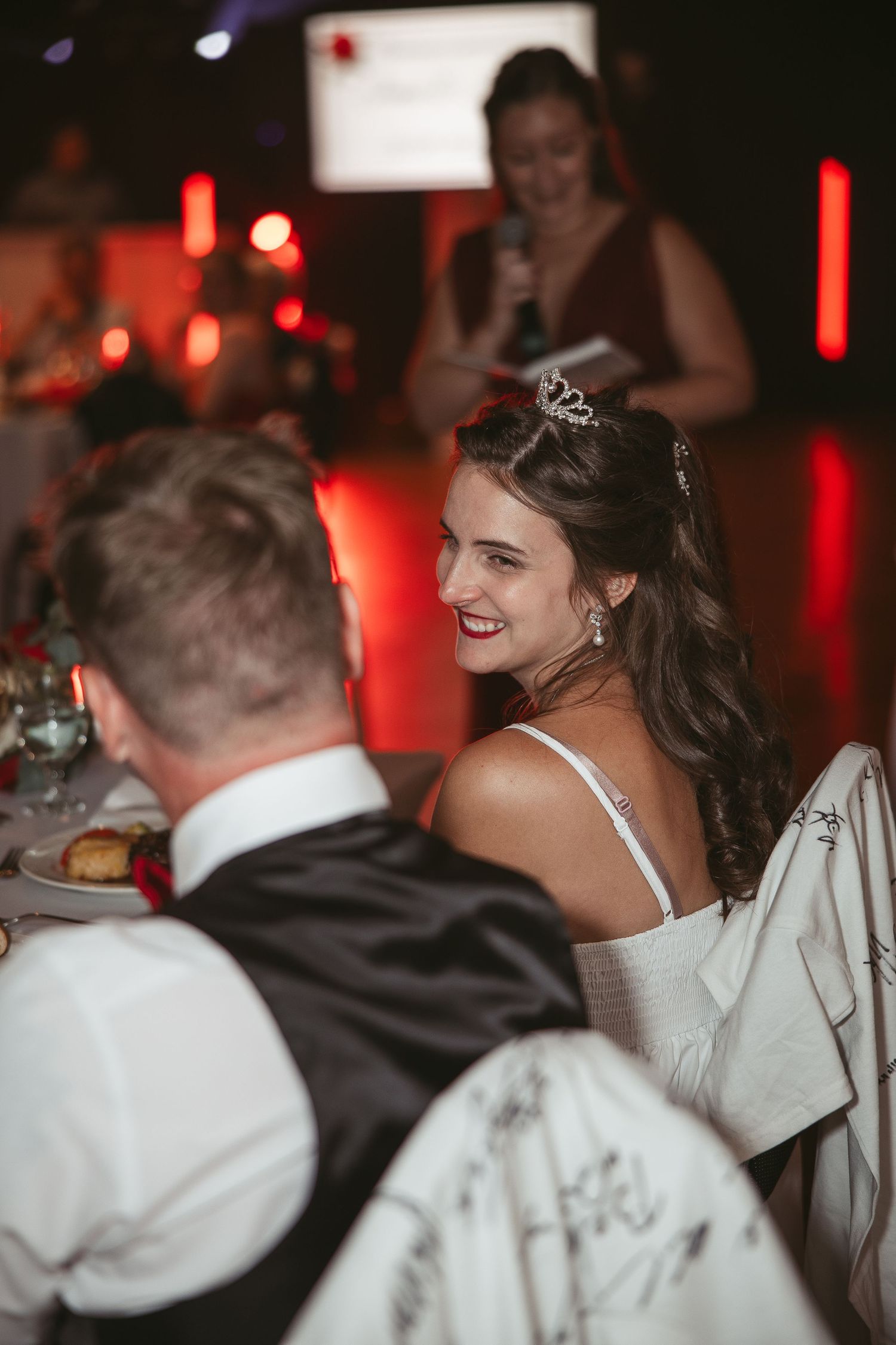La marié assise à la table d'honneur qui sourit en regardant son mari lors de la réception du mariage en août