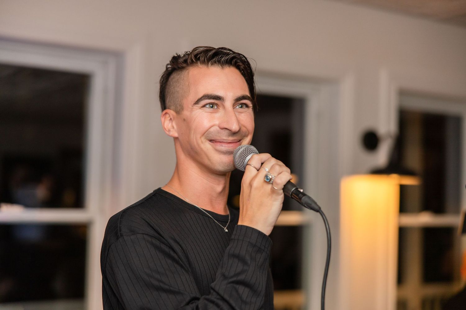 A person speaking into a microphone in a dimly lit room with windows in the background.
