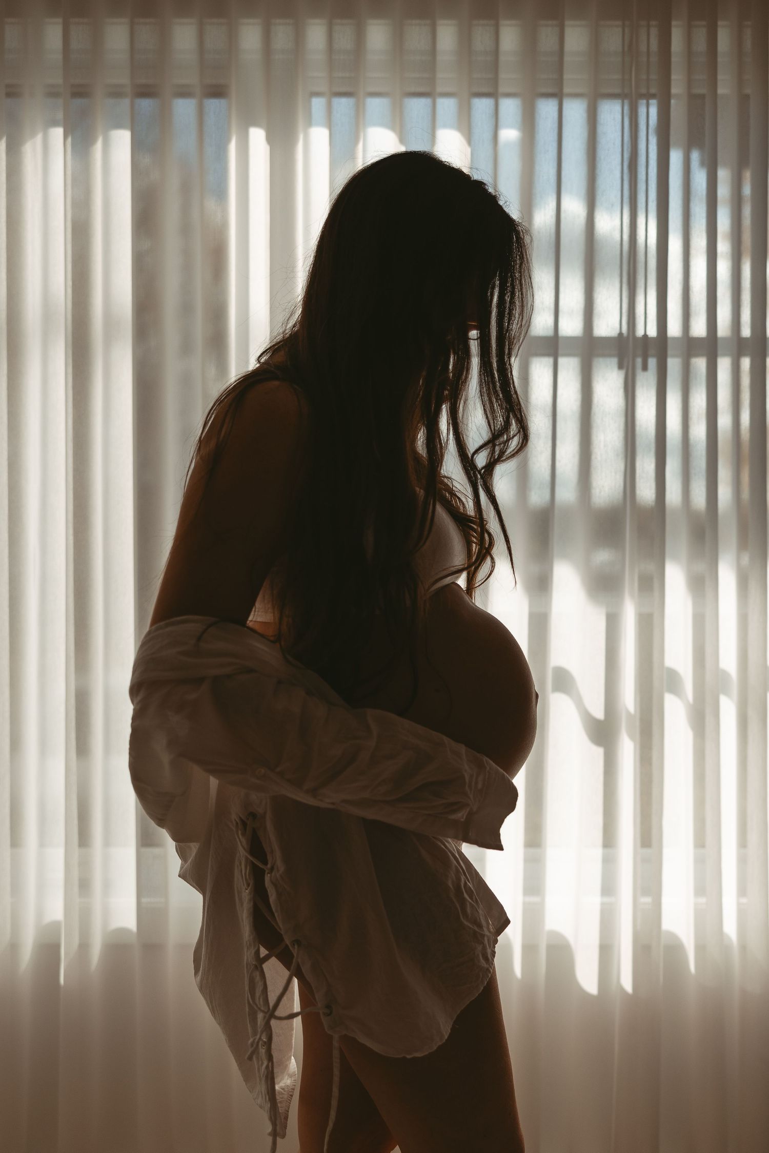 Femme enceinte en contre-jour devant fenêtre avec stores verticaux, silhouette capturée pour moments intimes et élégance de la maternité