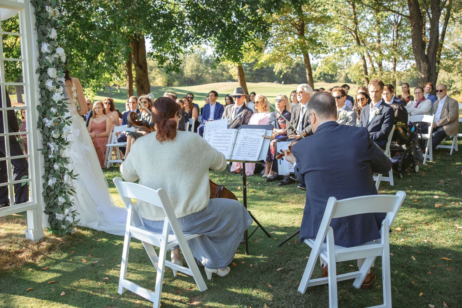 Guests seated in white chairs during an outdoor garden wedding ceremony under trees on a sunny afternoon.