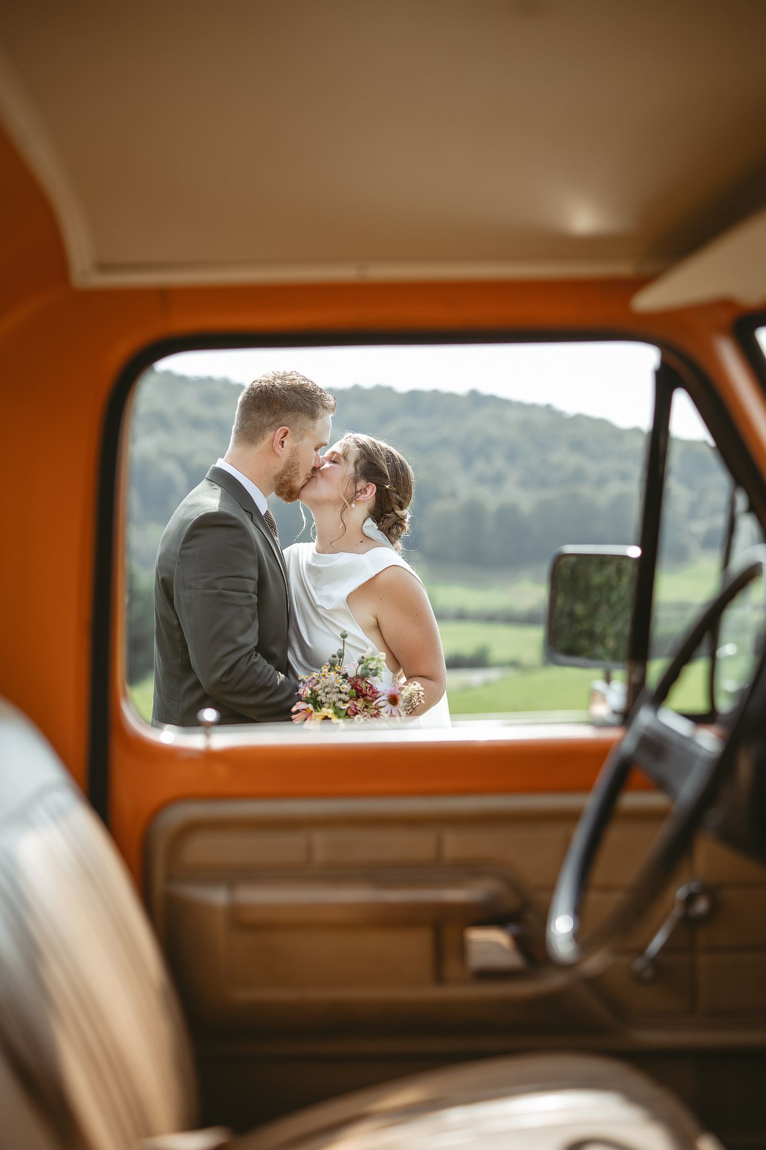 Couple de jeunes mariés partageant un moment intime vu à travers les vitres d’une voiture vintage, en décor champêtre