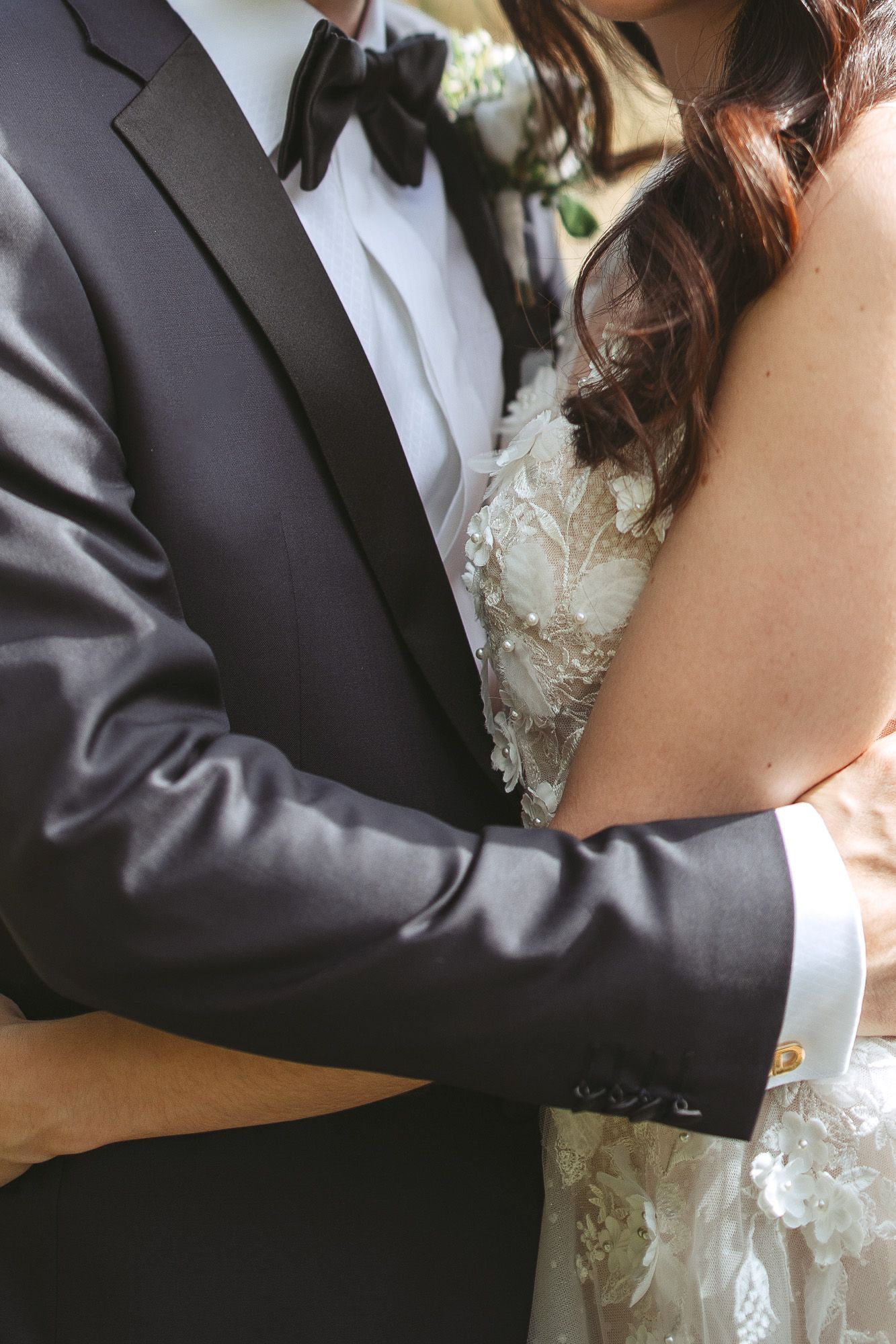 Gros plan d’un couple enlacé lors d’un mariage, détails d’un tuxedo noir et d’une robe blanche en dentelle